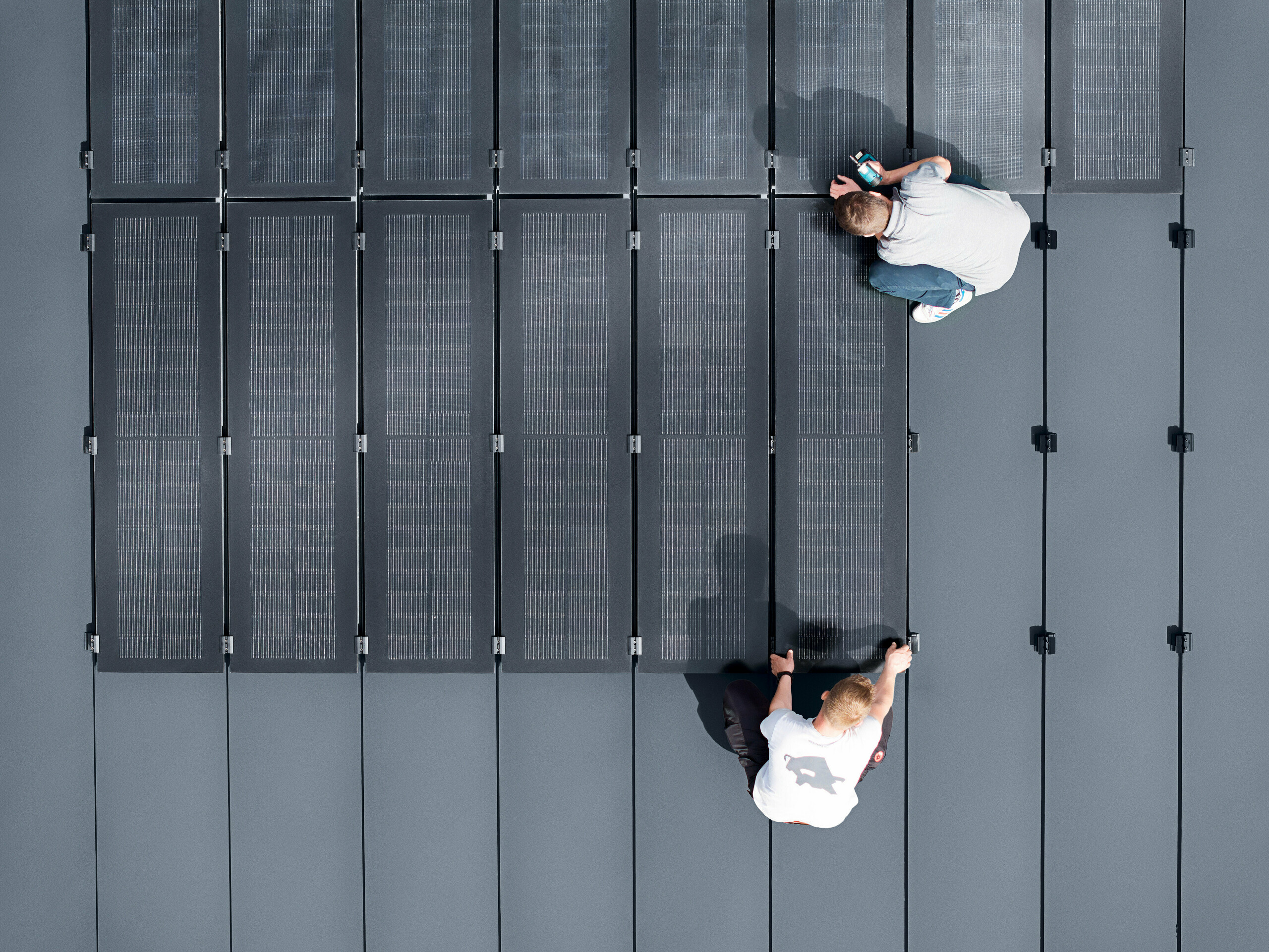 Zwei Personen installieren PREFALZ Solarmodule auf einer großen grauen Dachfläche, die mit Doppelstehfalzbahnen ausgelegt ist.