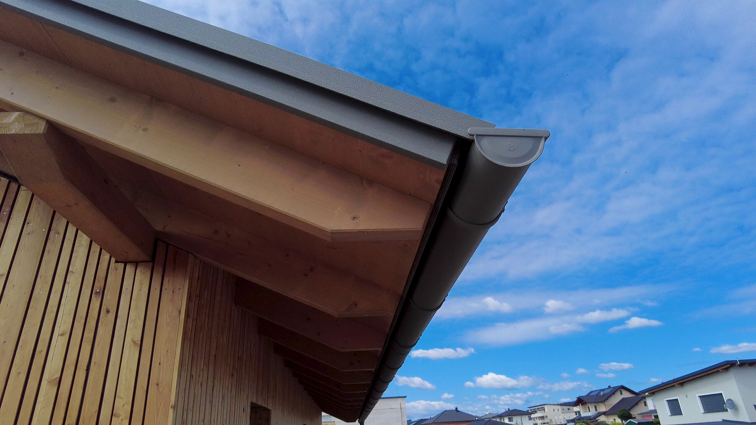 Der Blick unter den Dachvorsprung eines modernen Einfamilienhauses zeigt eine Holzverkleidung und eine Dachrinne von PREFA. Die sichtbaren Balken der Unterkonstruktion sowie die Dachrinne und das Abflussrohr sind in einem passenden Grauton gehalten. Das Foto ist an einem Tag mit leicht bewölktem blauem Himmel aufgenommen.