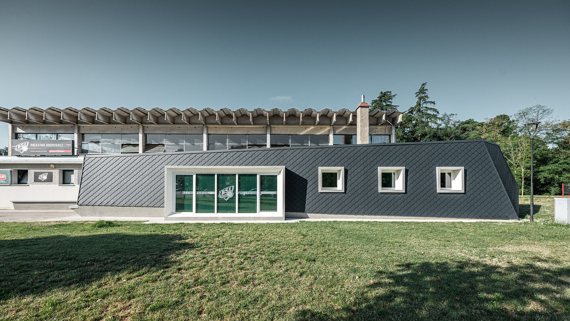 Ansicht einer modernen Turnhalle in Bologna, Italien, mit einer Fassade aus anthrazitfarbenen, rautenförmigen Aluminiumplatten von PREFA in 20x20 cm Format, vor einem bewaldeten Hintergrund und unter klarem Himmel.
