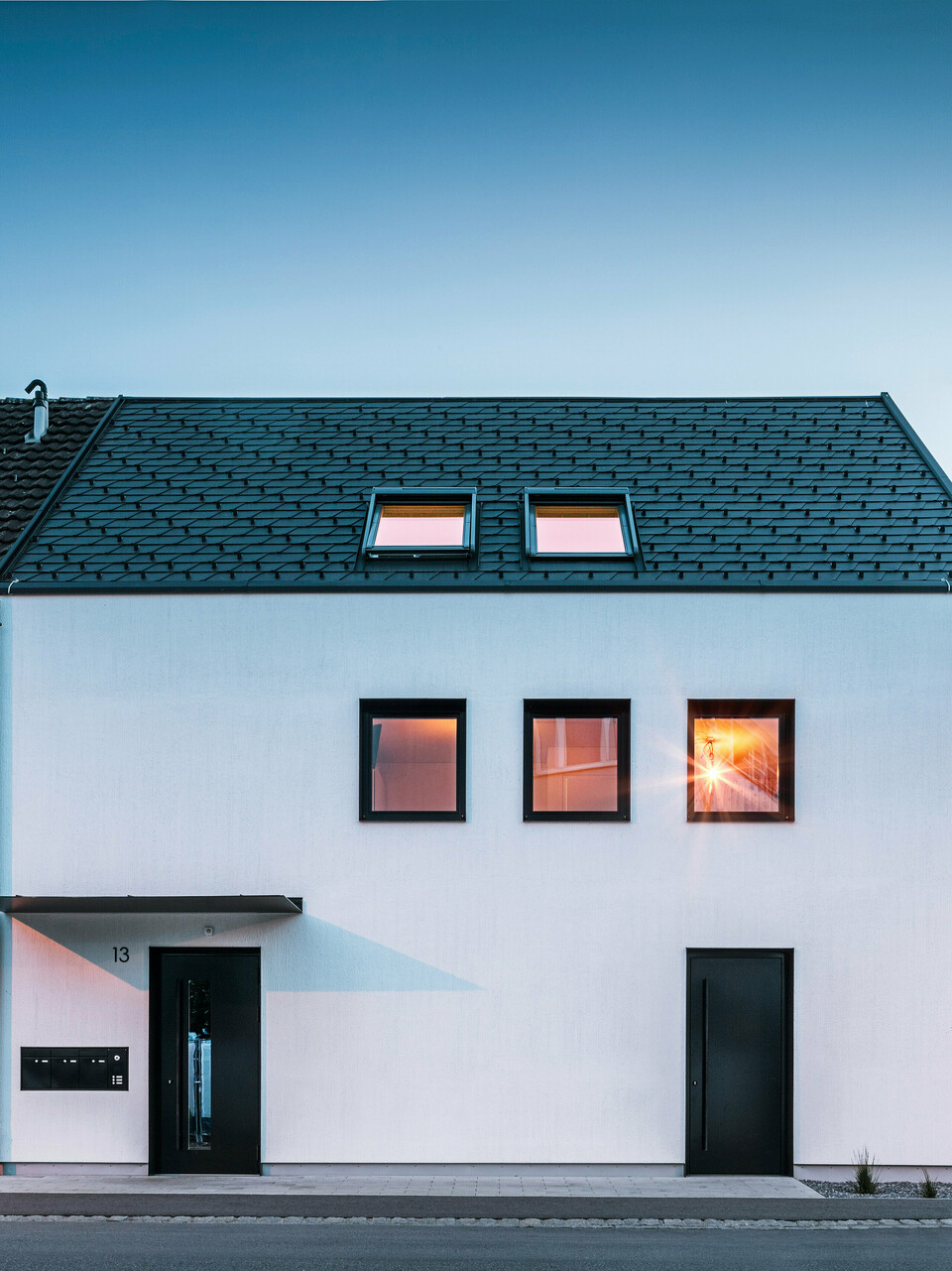 Moderne Mehrfamilienhausfassade mit PREFA Dachschindeln DS19 und Prefalz in Schwarz am Bodensee in Österreich. Das Gebäude hat eine klare Zweiteilung mit einem hölzernen Anbau auf der linken Seite und einem verputzten Hauptteil auf der rechten Seite. Das Dach zeigt die charakteristische dunkle Schindeldeckung und es gibt mehrere rechteckige Fenster, die im warmen Abendlicht spiegeln.
