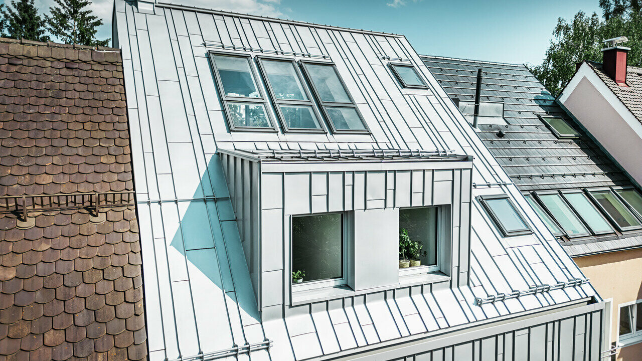 Ein modernes Reihenhaus mit einer Dach- und Fassadensanierung, die PREFA Aluminiumprodukte zeigt. Das Dach besteht aus silbermetallischen, kleinformatigen, stehfalzartigen Aluminiumplatten, die eine strukturierte, matte Oberfläche aufweisen und das Licht reflektieren. Die Hausfassade ist ebenfalls mit vertikalen Aluminiumelementen verkleidet, die einen zeitgemäßen Kontrast zu den traditionellen, terrakottafarbenen Ziegeldächern der angrenzenden Häuser bieten.