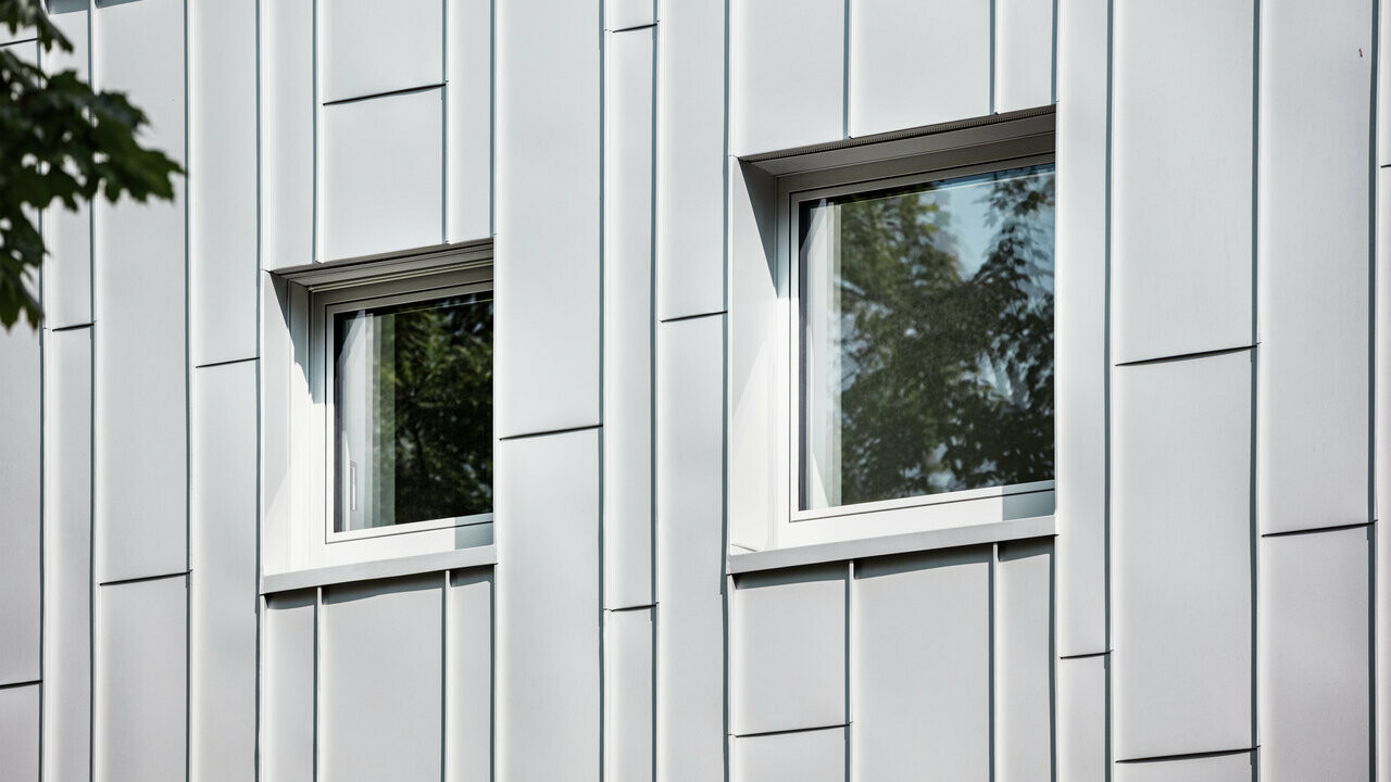 Nahaufnahme einer modernen Gebäudefassade mit vertikalen silbermetallischen Prefalz Aluminiumpaneelen. Zwei rechteckige Fenster mit dunklen Rahmen spiegeln das Grün der Bäume wider und durchbrechen das strukturierte Fassadenmuster.