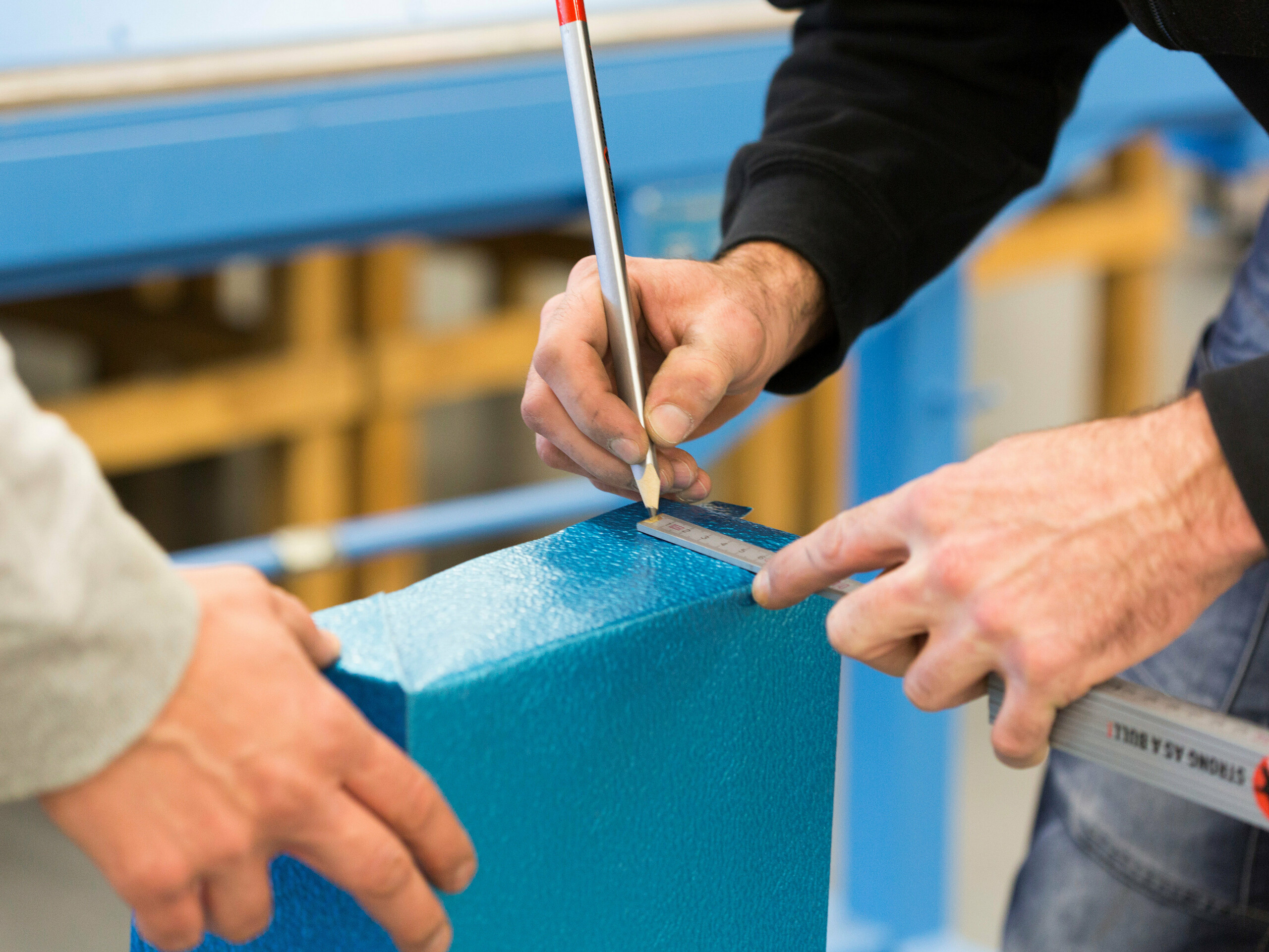 Zwei Handwerker bei der präzisen Markierung eines PREFA Produktes aus blauem Aluminium. Die Männerhände werden unterstützt durch professionelle Werkzeuge und Materialien. Der Maßstab in der Hand des zeichnenden Mannes trägt ein rotes PREFA Logo. Das Bild symbolisiert die Unterstützung von Spenglern durch die Firma PREFA. 