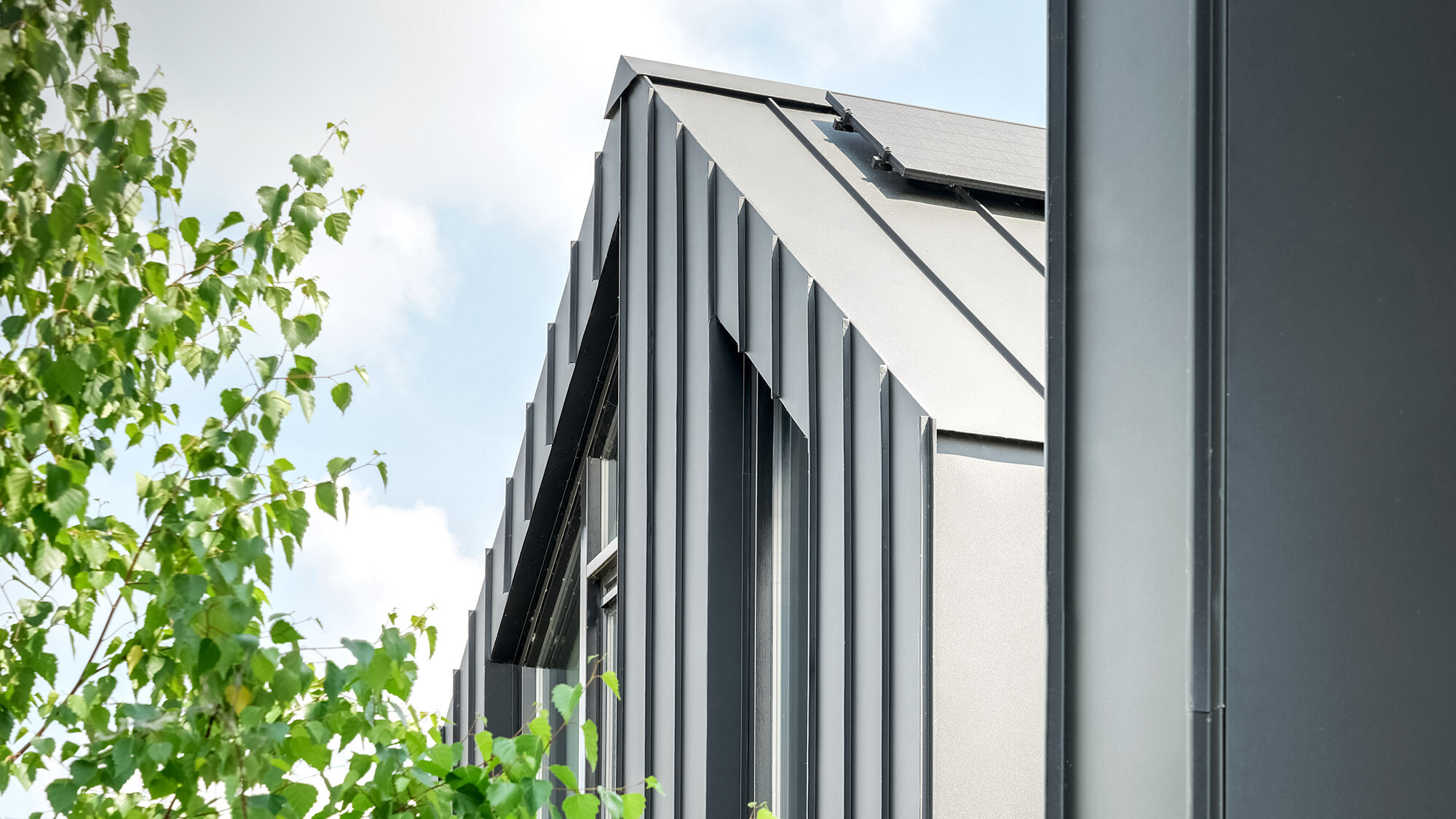 Seitenansicht eines modernen Hauses mit einem steilen Satteldach, gedeckt mit schwarzen Prefalz-Dachbahnen, und ausgestattet mit Solarmodulen. Die vertikalen Fassadenelemente und die dunkle Dachfarbe kontrastieren mit der lebendigen grünen Umgebung aus nahen Baumzweigen, die in das Bild hineinragen.