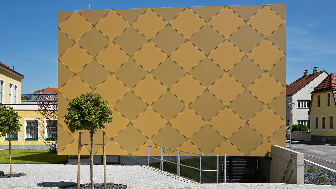 Eingangsbereich des Musikvereins Zwettl. Das markante Gebäude in Quaderform ist umhüllt von PREFA Aluminium Verbundplatten. Die karoförmigen Aluminiumplatten in zwei unterschiedlichen Goldtönen lassen das Objekt erstrahlen.