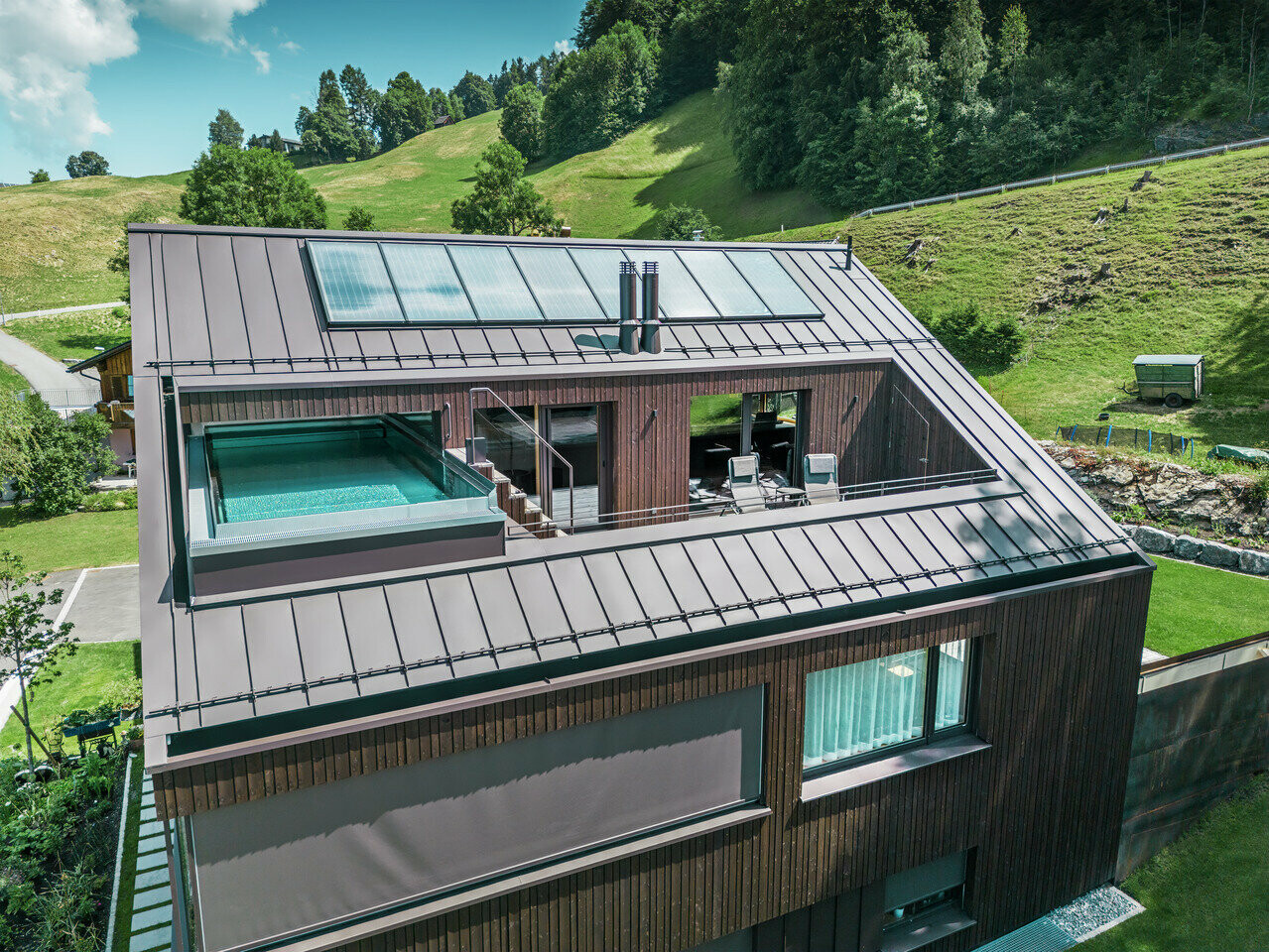 Ansicht eines modernen Mehrfamilienhauses mit einem PREFA Dach in Prefalz-Technik, ausgeführt in der Farbe Nussbraun. Das Bild zeigt auch eine Terrasse mit einem Pool, umgeben von einer Holzverkleidung und Glasgeländern.