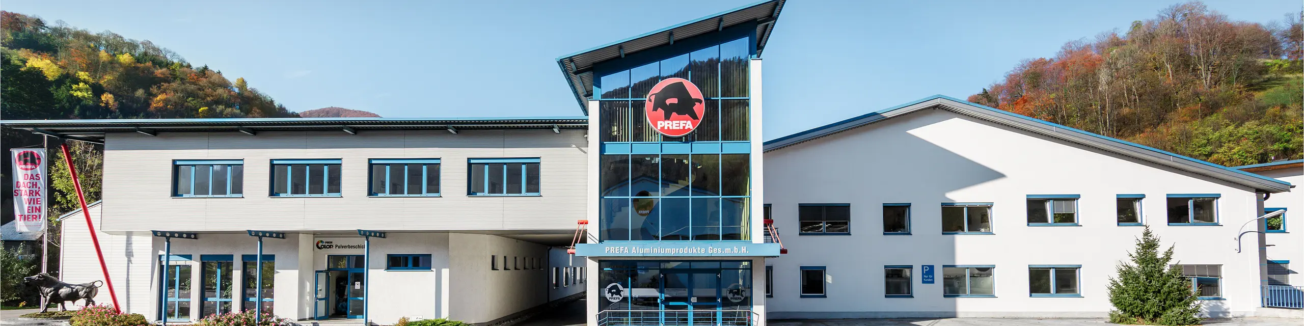 Markante Frontansicht des PREFA Standorts Marktl mit großem PREFA-Stier-Logo, moderner Glasfassade und klarer Architektur vor herbstlicher Hügellandschaft. Das Gebäude steht für die Qualität und Innovationskraft von PREFA Aluminium. 