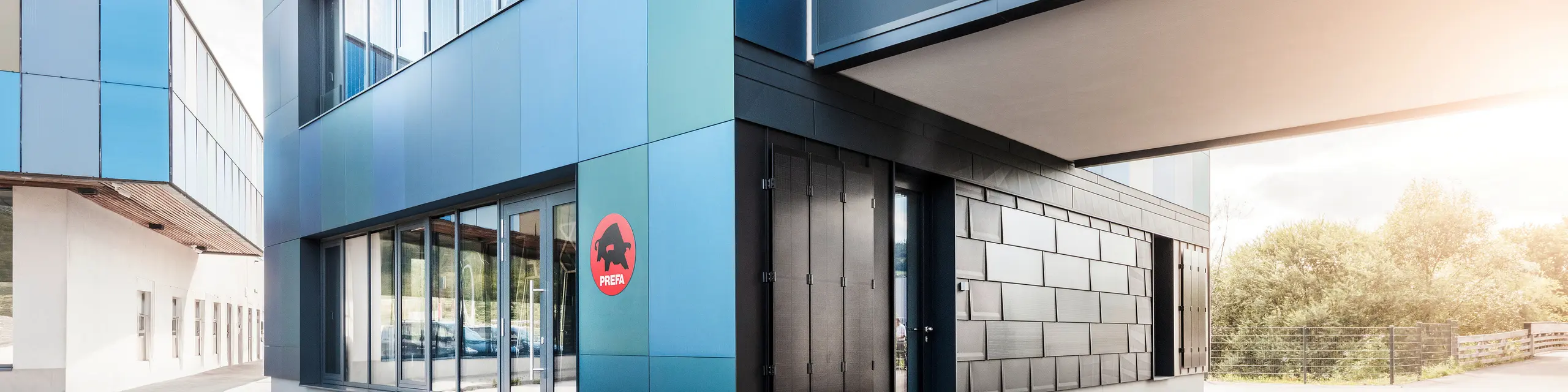Außenansicht des PREFA Showrooms in St. Veit mit modernen Fassadenplatten in einer Farbmischung aus Blau- und Grüntönen. Das Gebäude verfügt über große Fenster und zeigt das PREFA Logo an der Fassade.