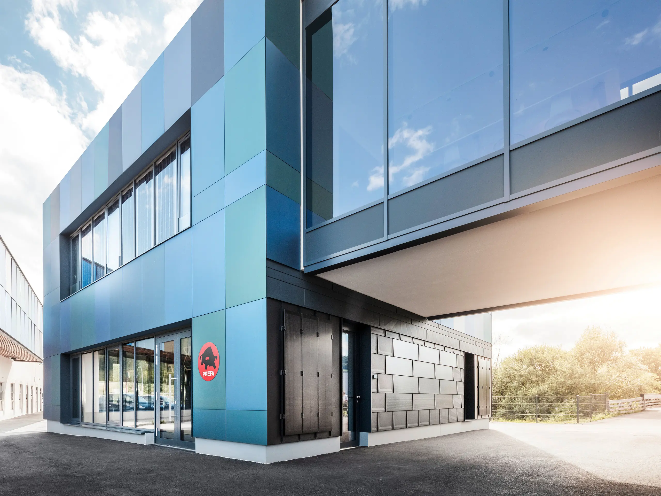 Außenansicht des PREFA Showrooms in St. Veit mit modernen Fassadenplatten in einer Farbmischung aus Blau- und Grüntönen. Das Gebäude verfügt über große Fenster und zeigt das PREFA Logo an der Fassade.