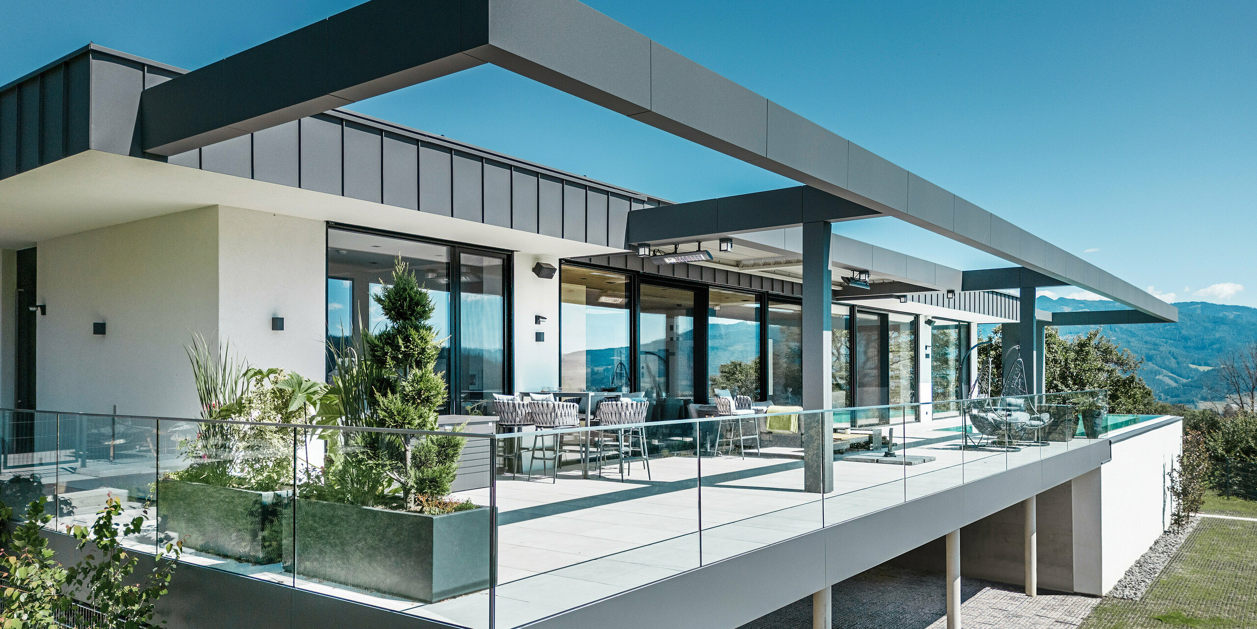 Luxuriöse Terrasse eines Einfamilienhauses in Pogier, Österreich, mit einem eleganten Dach- und Fassadensystem von PREFA. Die Kombination aus PREFALZ und PREFABOND in Dunkelgrau schafft ein modernes und anspruchsvolles Design. Die großzügige Terrasse bietet eine atemberaubende Aussicht und ist mit Glasgeländern ausgestattet, die für eine offene und einladende Atmosphäre sorgen. Die teilweise überdachte Außenfläche ermöglicht es, die Schönheit der umliegenden Natur zu genießen und ist ideal für Entspannung und soziale Zusammenkünfte.