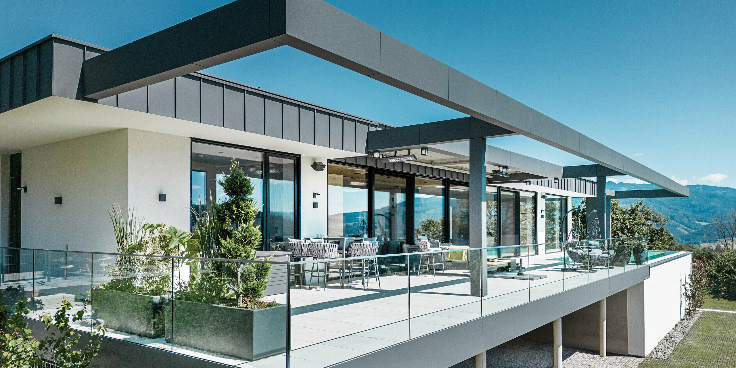 Luxuriöse Terrasse eines Einfamilienhauses in Pogier, Österreich mit eleganten Dach- und Fassadensystemen von PREFA. Die Kombination von PREFALZ und PREFABOND in dunkelgrau ergibt ein modernes und anspruchsvolles Design. Die großzügige Terrasse bietet einen atemberaubenden Ausblick und ist mit einem Glasgeländer ausgestattet, das eine offene und einladende Atmosphäre schafft. Der teilweise überdachte Außenbereich lässt die Schönheit der umliegenden Natur genießen und ist ideal zum Entspannen und geselligen Beisammensein.