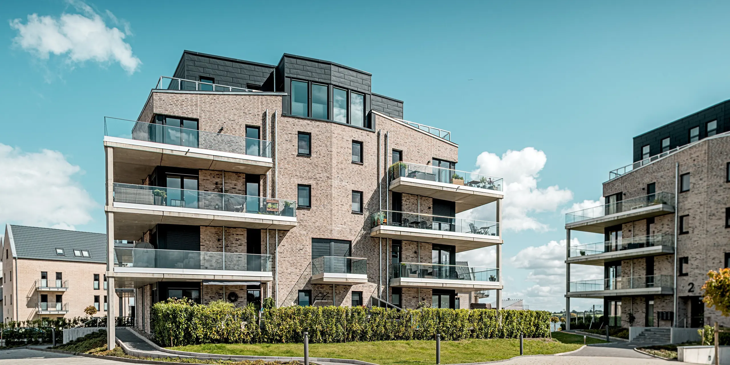 Die Wohnhausanlage „Hafen zur Freiheit“ präsentiert sich in einer offenen Hofsituation, bei der sich moderne Klinkerarchitektur harmonisch mit den markanten PREFA Siding.X Akzenten in P.10 Anthrazit verbindet. Das zentrale Gebäude mit seinen auskragenden Balkonen und klaren Linien steht im Mittelpunkt und wirkt durch die dynamisch strukturierten Aluminiumflächen besonders kraftvoll. Großzügige Glasbrüstungen verstärken den modernen Look und reflektieren das Tageslicht. Die langlebigen, korrosionsbeständigen PREFA Fassadenpaneele bieten optimalen Schutz vor den rauen Küstenbedingungen und unterstreichen die hochwertige Bauqualität.