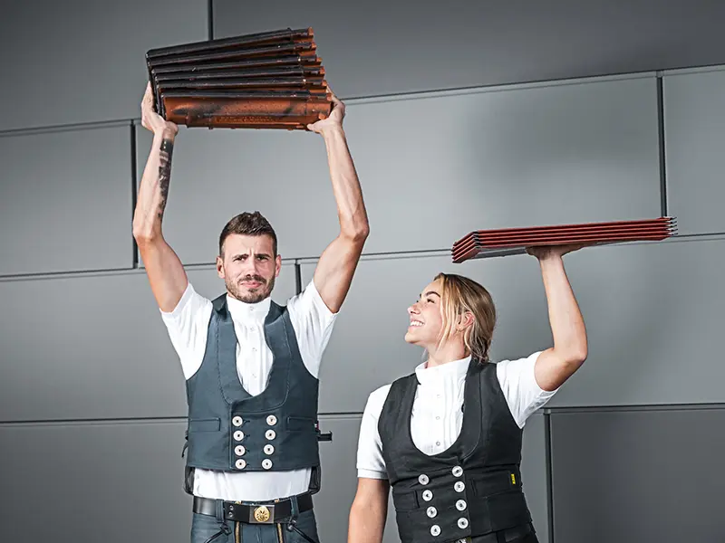 In dem Bild sind zwei Personen zu sehen, die jeweils ein Stapel Dachdeckmaterial in der Hand halten. Die Person auf der linken Seite hält einen Stapel Dachziegel über dem Kopf, während die Person auf der rechten Seite einen Stapel Aluminiumdachplatten ebenfalls über dem Kopf hält. Beide tragen ein traditionelles Handwerker-Outfits mit einer Weste und einem weißen Hemd. Der Hintergrund ist modern und minimalistisch, mit grauen Wänden. Beide Personen wirken konzentriert und lächeln.