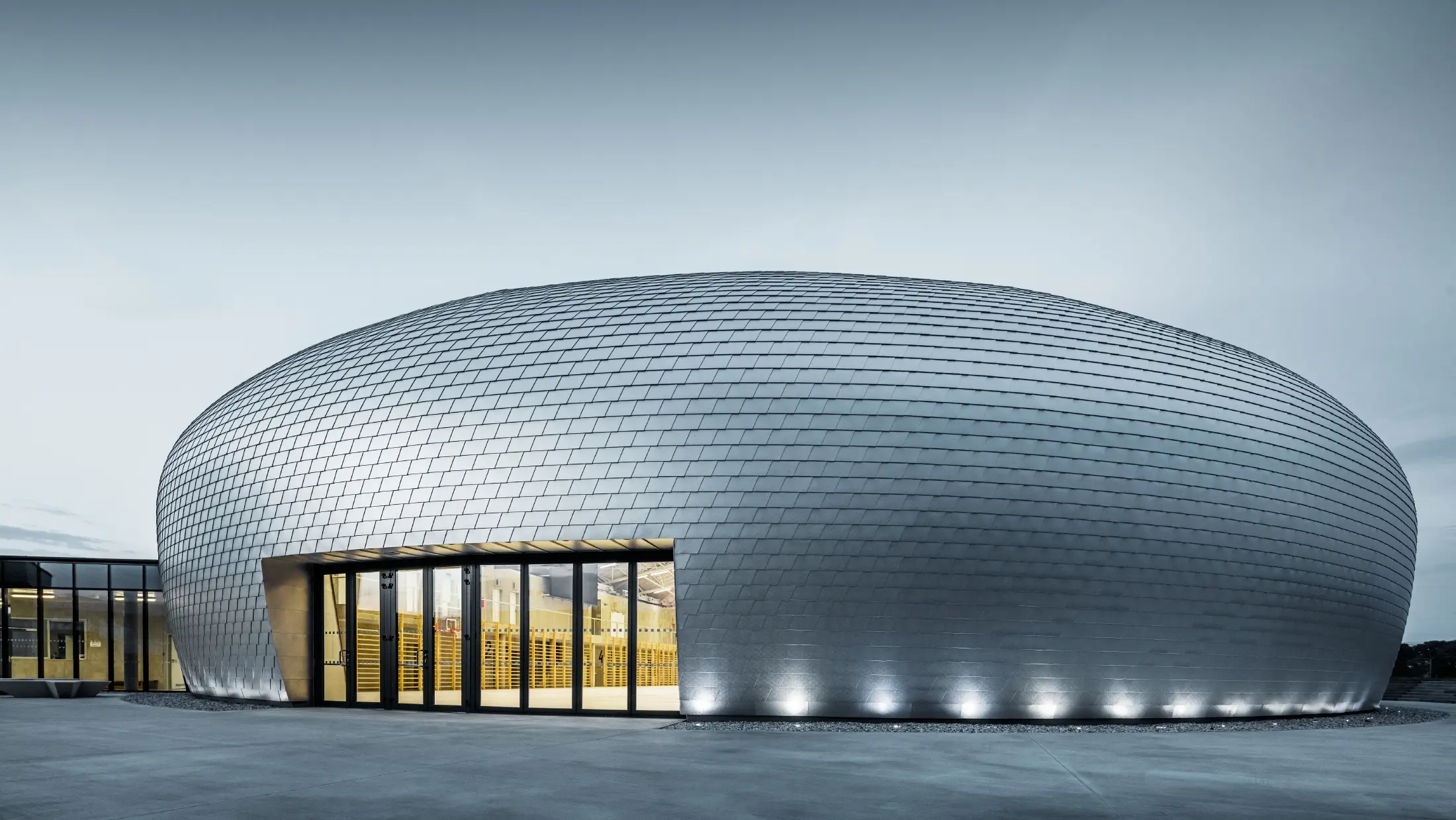 Sporthalle in Tschechien in Form eines UFOs verkleidet mit der PREFA Wandschindel in Naturblank bei Sonnenaufgang.