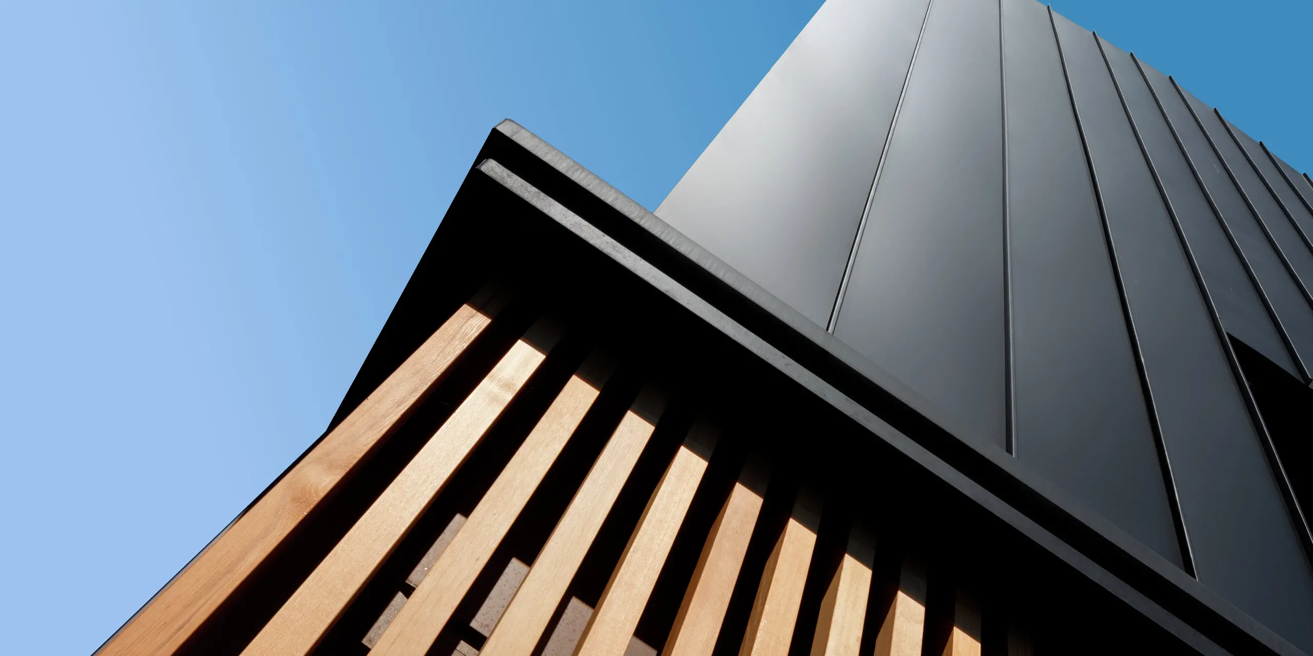 Architekturdetail eines modernen Doppelhauses in Cattolica: vertikale Aluminiumverkleidung mit PREFALZ in Schwarzgrau trifft auf elegante Holzlamellen. Die klare Linienführung und die hochwertige PREFA Oberfläche sorgen für Langlebigkeit, Korrosionsschutz und ein markantes Design unter strahlend blauem Himmel.