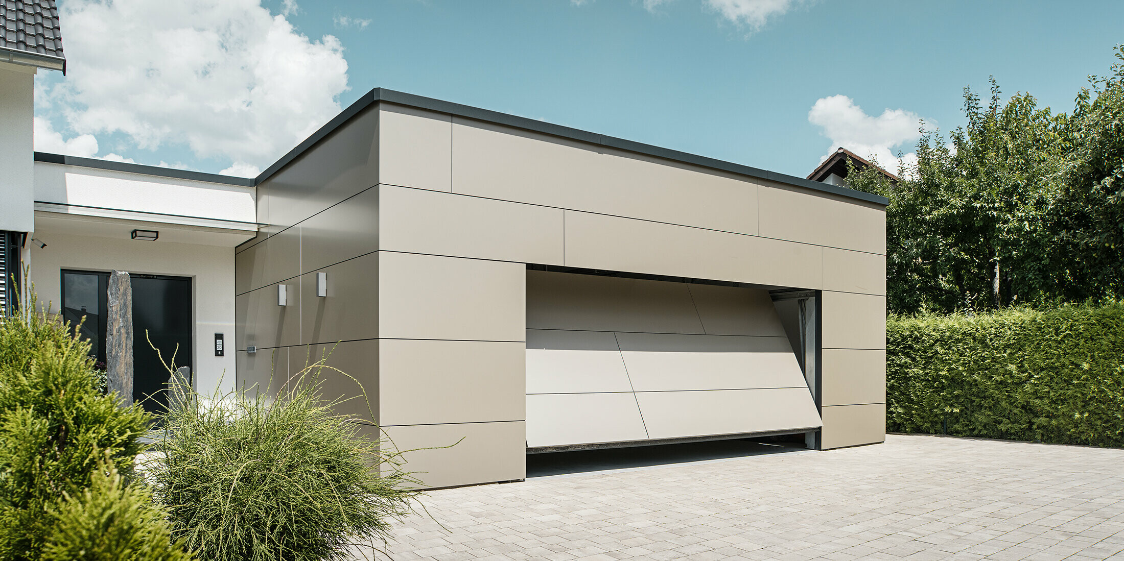 Gesamtansicht einer mit PREFABOND Aluminium Verbundplatten in Bronze verkleideten Garage. Das automatische Garagentor öffnet sich gerade und ist bereits ein Stück geöffnet. Direkt neben der Garage befindet sich der Hauseingang mit anthrazitfarbener Tür und weißer Putzfassade.