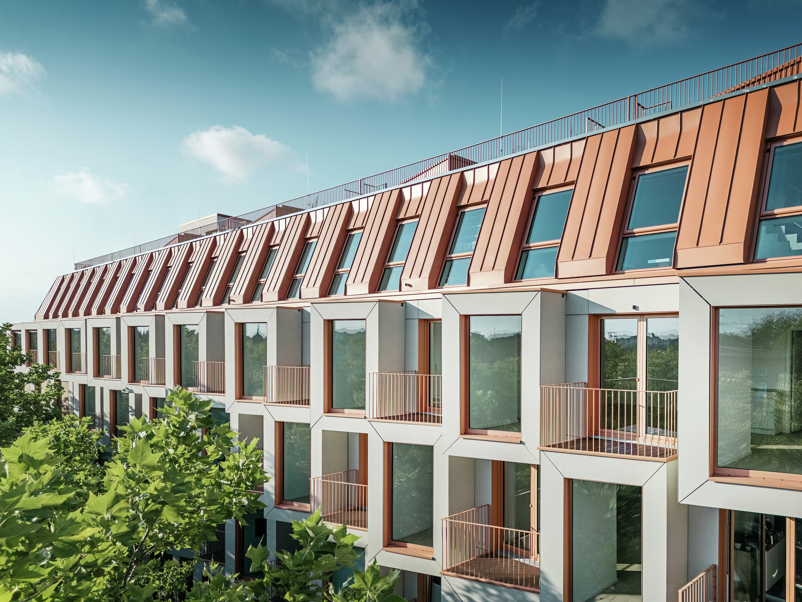 Fassadenansicht des Wohnbauprojektes Van B in München mit dem markanten PREFALZ Aluminiumdach in der Sonderfarbe IGP Orange HDP von PREFA. Die moderne Dacheindeckung verbindet ästhetische Eleganz mit technischer Präzision und bietet dauerhaften Schutz vor Witterungseinflüssen. Die kubisch gestalteten Erker mit großzügigen Fensterflächen und filigranen Balkonen schaffen ein harmonisches Zusammenspiel von Licht, Raum und Funktionalität - ideal für anspruchsvolle Wohnkonzepte im urbanen Umfeld.