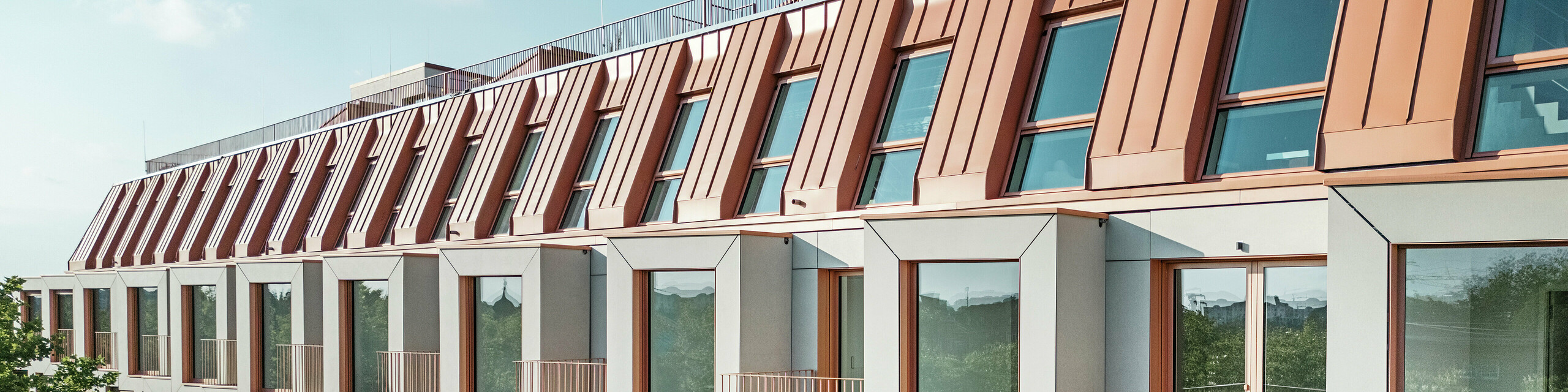 Fassadenansicht des Wohnbauprojektes Van B in München mit dem markanten PREFALZ Aluminiumdach in der Sonderfarbe IGP Orange HDP von PREFA. Die moderne Dacheindeckung verbindet ästhetische Eleganz mit technischer Präzision und bietet dauerhaften Schutz vor Witterungseinflüssen. Die kubisch gestalteten Erker mit großzügigen Fensterflächen und filigranen Balkonen schaffen ein harmonisches Zusammenspiel von Licht, Raum und Funktionalität - ideal für anspruchsvolle Wohnkonzepte im urbanen Umfeld.