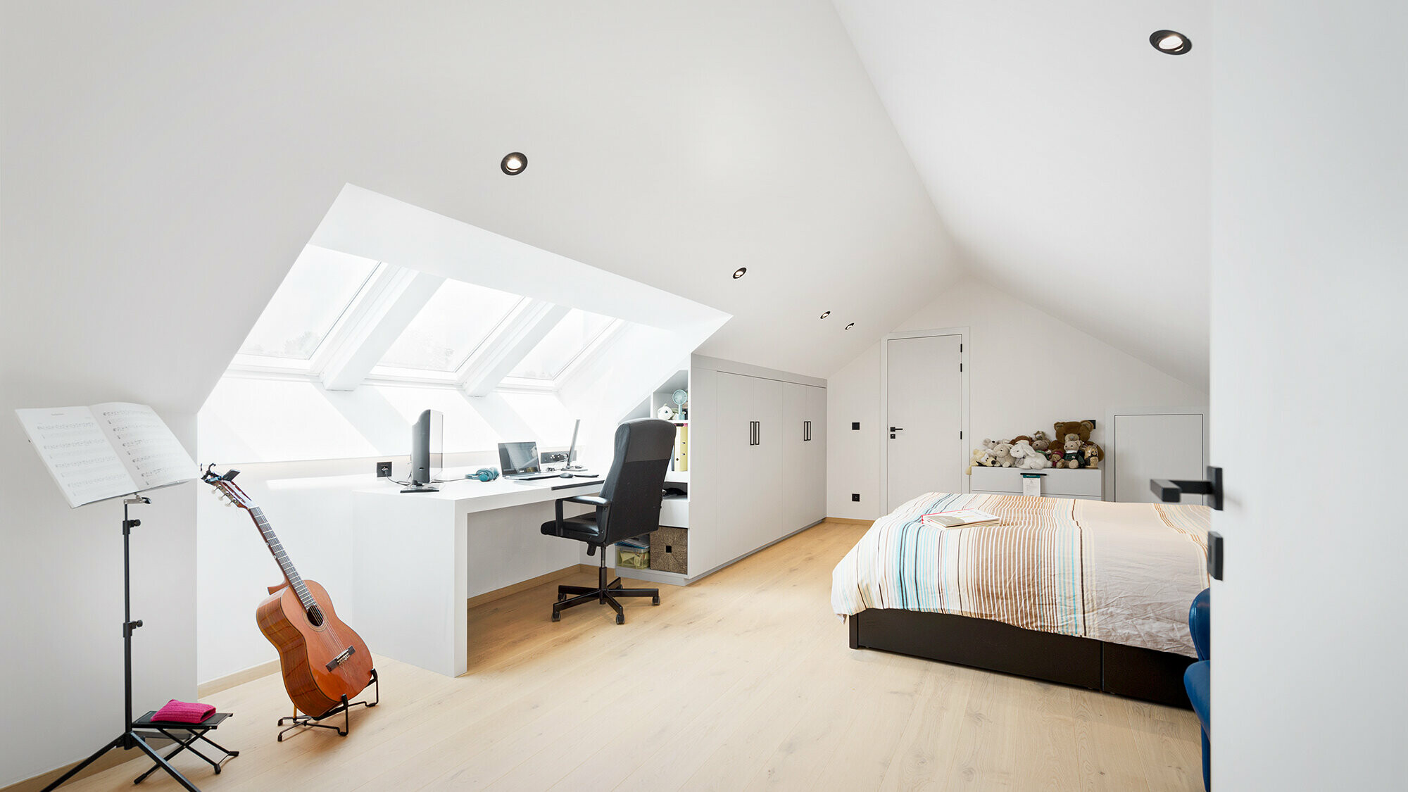 Innenansicht einer der hellen Zimmer im Dachgeschoss samt Bett, Schreibtisch, Gitarre und Notenständer.