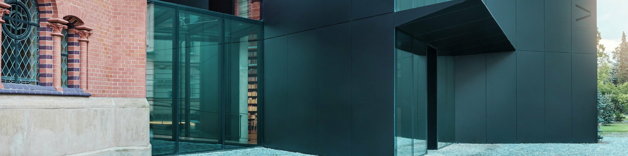 Das Bild zeigt die moderne Erweiterung einer Bibliothek, die sich elegant an die historische Rote Kirche in Olmütz anschließt. Der markante Neubau beeindruckt mit einer Fassade aus schwarzen PREFABOND Aluminium-Verbundplatten, die in klaren, geometrischen Formen gestaltet ist und mit präziser Detailarbeit überzeugt. Die Glaswände des Gebäudes bieten einen transparenten Blick in das Innere, das sich harmonisch in die historische Umgebung einfügt. Das Zusammenspiel von modernen Materialien und klassischen Architekturelementen schafft eine spannende visuelle Verbindung zwischen Alt und Neu. Der innovative Einsatz der PREFABOND Platten sorgt nicht nur für ein modernes Erscheinungsbild, sondern auch für Langlebigkeit und Widerstandsfähigkeit, die das Gebäude für die Zukunft wappnen.