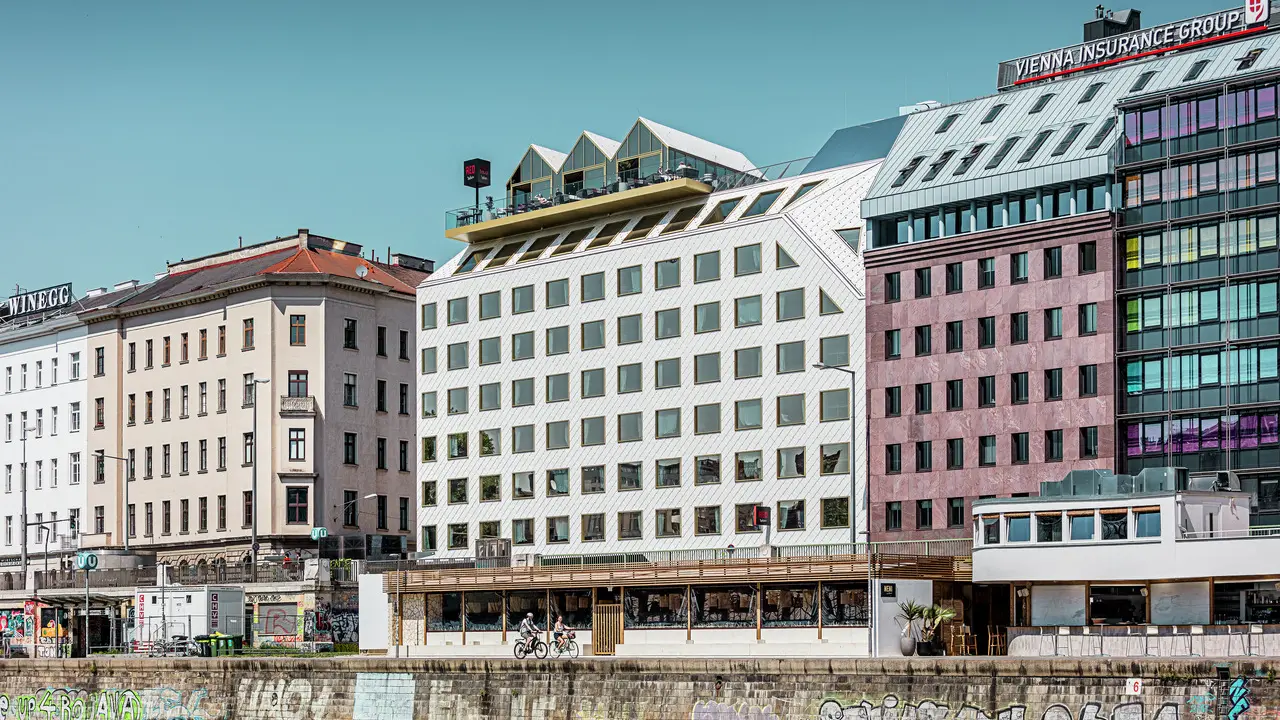 Blick auf das Ufer des Donaukanals in Wien mit einer Reihe von Gebäuden unterschiedlicher Architektur. Im Vordergrund ist das "THE ROCK Radisson RED Vienna" Hotel, erkennbar an seiner charakteristischen weißen PREFA Aluminiumfassade mit Dach- und Wandrauten 44 × 44 in P.10 Reinweiß.