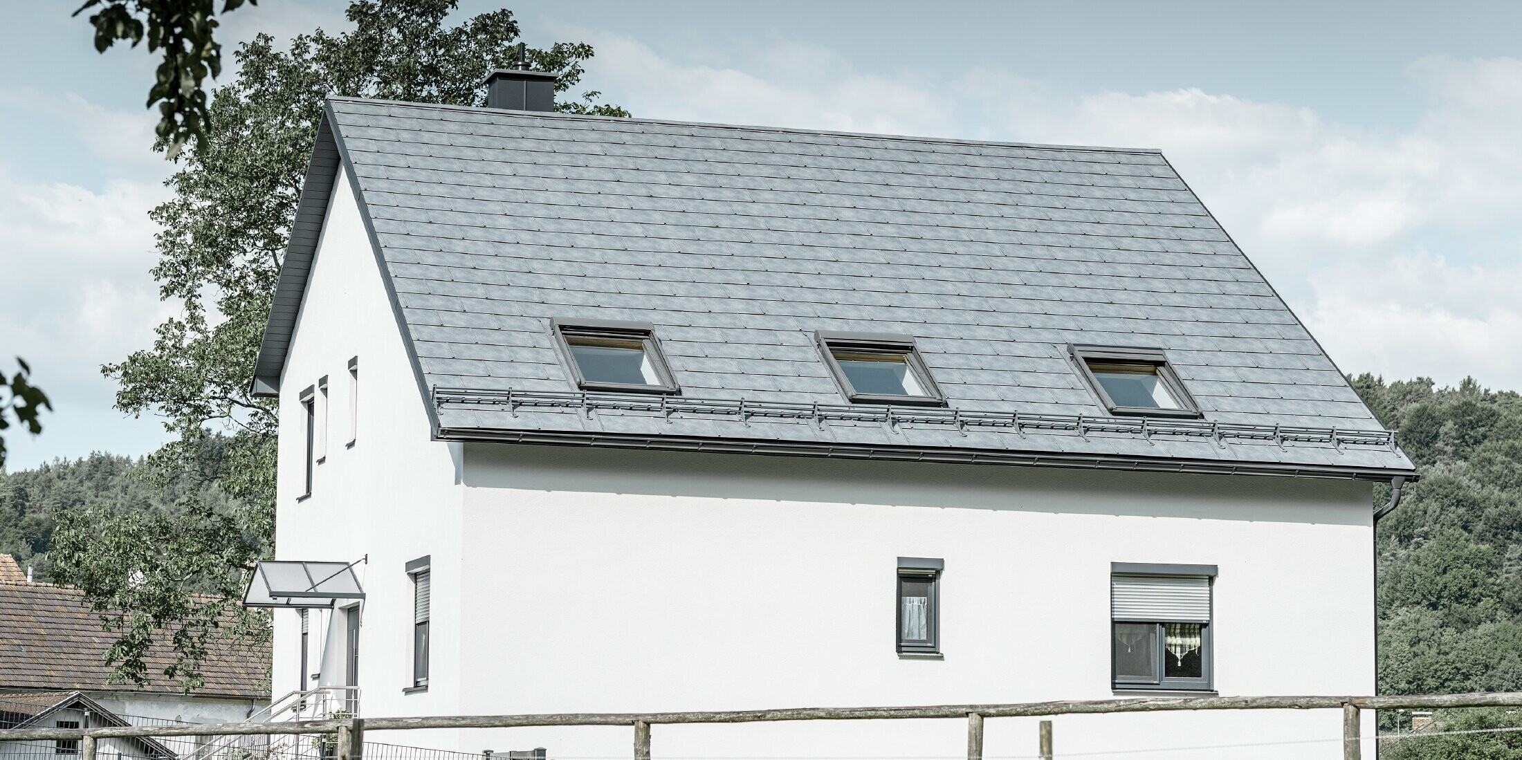 Das Satteldach des klassischen Einfamilienhauses wurde mit der neuen PREFA Dachplatte R.16 in Steingrau eingedeckt. In der Dachfläche wurden drei Dachfenster eingefügt und Schneerechen montiert. Die Fassade ist einfach in Weiß gehalten.