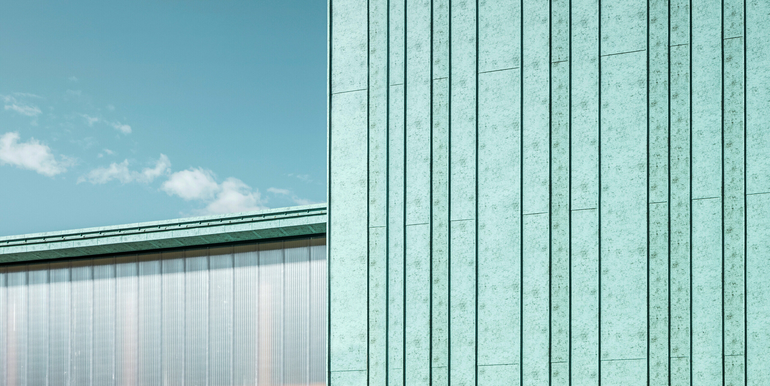 Das Bild zeigt eine moderne PREFA Fassade aus Aluminium in einem einzigartigen patinagrünen Farbton. Die Fassade präsentiert sich in einer vertikalen Stehfalzdeckung, was ihr eine strukturierte Ästhetik verleiht. Die klaren Linien der vertikalen Fassadenelemente bieten einen harmonischen Kontrast zum blauen Himmel und der natürlichen Umgebung im Hintergrund. Bei dem Gebäude handelt es sich um das Zero Magnetic Field Laboratory in Fertöboz (HU). Das Observatorium ist gut in seine Umgebung integriert, wobei die Farbe der Fassade eine ökologische und naturnahe Anmutung hat. Die Fassade wirkt trotz ihrer sichtbaren modernen Beschaffenheit und des industriellen Materials natürlich und fügt sich sanft in die Landschaft ein.