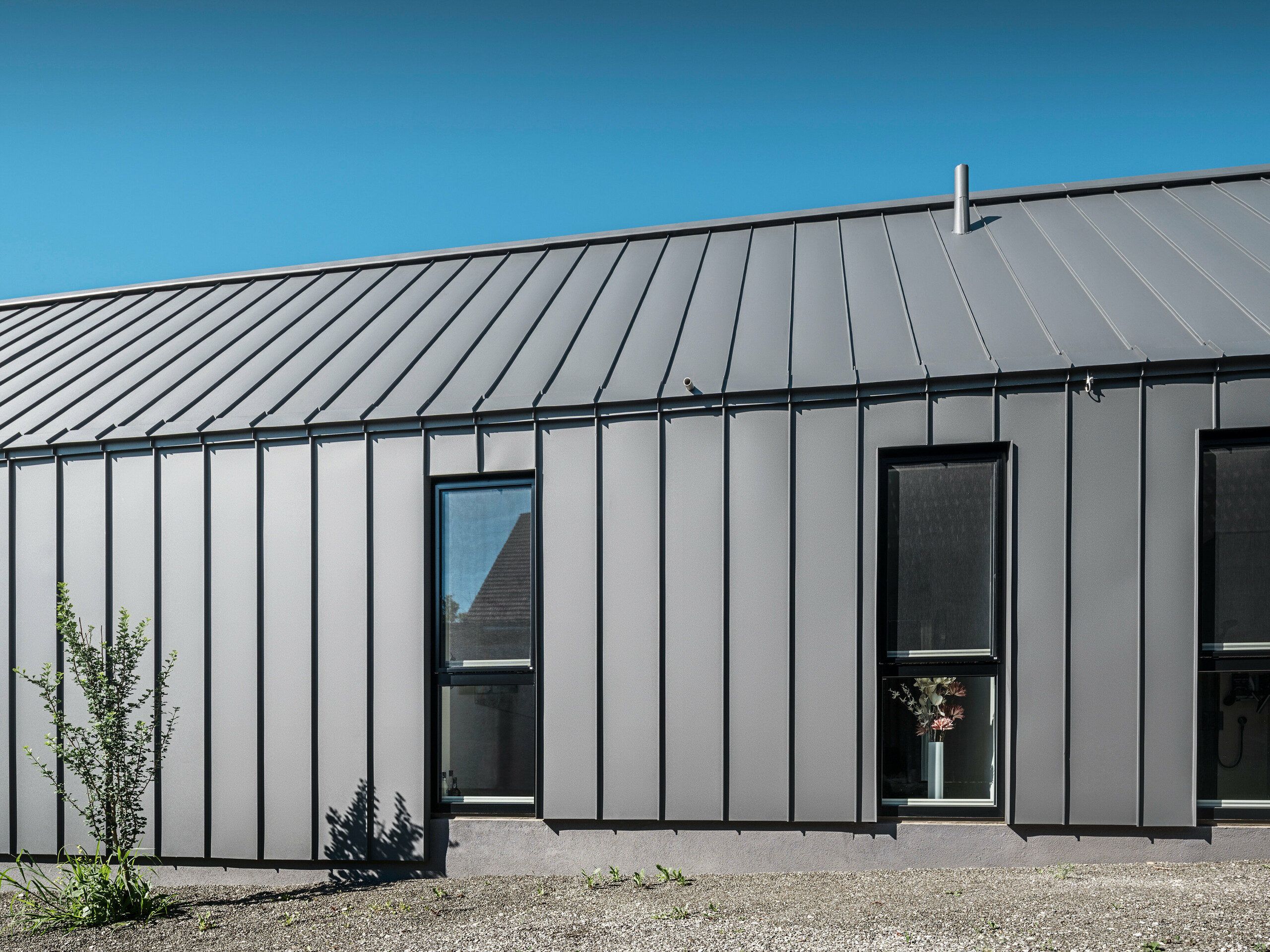 Seitenansicht eines Einfamilienhauses in Pogier, Österreich, mit langlebigem und elegantem PREFALZ in P.10 dunkelgrau an Dach und Fassade. Die Aluminium-Stehfalzverkleidung bietet eine klare vertikale Linienführung, die die Fenster elegant umrahmt und für ein modernes Erscheinungsbild sorgt. Die minimalistische Architektur steht unter einem klaren blauen Himmel.