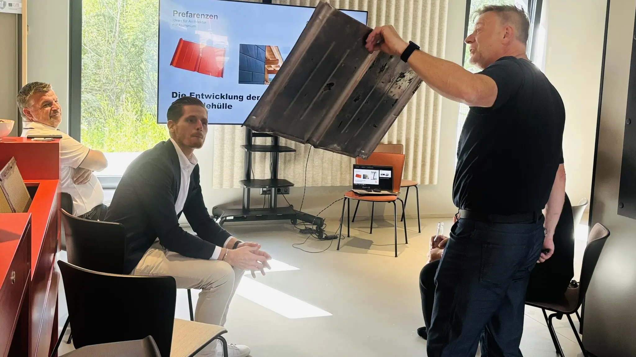 Im PREFA Showroom St. Veit an der Glan wird im Rahmen des Barcamp-Abends eine Aluminium-Dachplatte praxisnah präsentiert. Ralf Stadlober, PREFA Objektberater in Kärnten und Osttirol, hält ein großformatiges Musterelement und erläutert die Eigenschaften im Zusammenspiel mit gezeigten Referenzen auf einem Bildschirm. Mehrere Teilnehmende sitzen und verfolgen die Demonstration aufmerksam. Im Hintergrund sind Präsentationsflächen, ein Monitor mit Architekturbeispielen sowie Musterlösungen zu sehen. Die Szene steht für interaktiven Wissenstransfer, praxisnahe Einblicke und die vielseitigen Einsatzmöglichkeiten von PREFA Aluminium für Dach und Fassade.