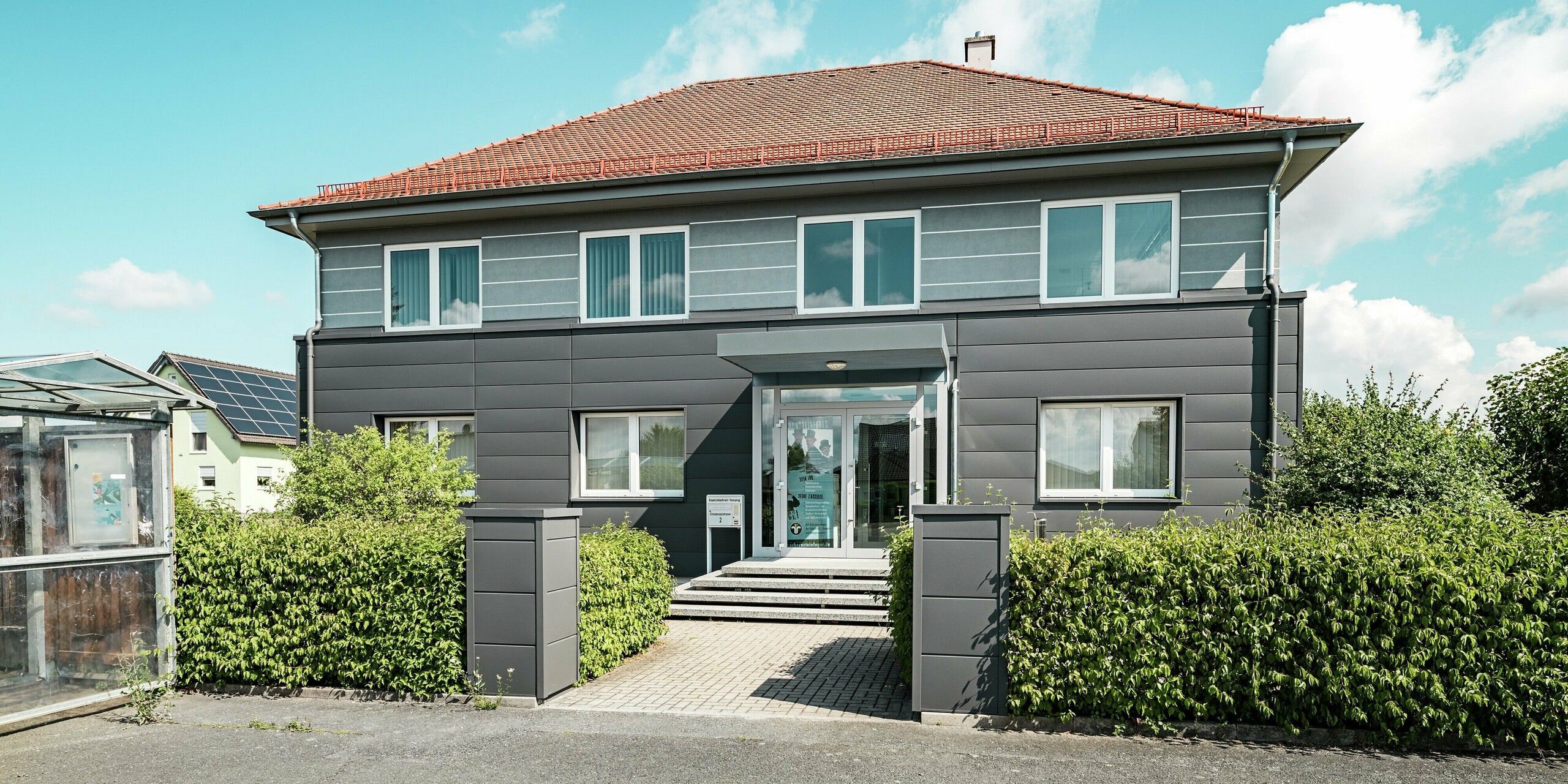 Die sanierte Fassade der Kaminkehrer-Innung in Bindlach ist mit hochwertigen PREFA Sidings in der Farbe P.10 Dunkelgrau verkleidet. Die klar strukturierten Aluminium-Paneele verleihen dem Gebäude eine zeitgemäße, elegante Optik. Der Eingang wird durch das moderne Design betont, während die Paneele entlang der Fassade nahtlos verlegt sind. Das Material ist nicht nur langlebig und pflegeleicht, sondern sorgt auch für nachhaltigen Schutz und fügt sich harmonisch in die Umgebung ein. Die grüne Bepflanzung und der klare Himmel bilden einen schönen Kontrast zum grauen Gebäude mit roten Ziegeln am Walmdach.