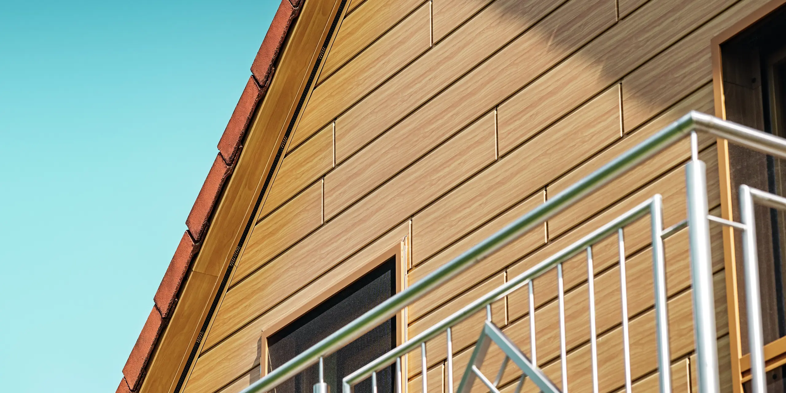 Detailaufnahme des sanierten Giebels eines Einfamilienhauses in Blieskastel. Der Giebel wurde mit PREFA Sidings in der Farbe Eiche Natur verkleidet, wodurch er eine warme, moderne Holzoptik erhält. Die feine Struktur und der natürliche Farbton der Aluminiumfassade sehen aus wie echtes Holz und setzen einen starken visuellen Akzent unter dem klaren Sommerhimmel. Gleichzeitig bleibt die Oberfläche dauerhaft witterungsbeständig, farbstabil und nahezu wartungsfrei. Sie ist ein langlebiges Highlight, das sich harmonisch in die Balkongeländer und das rote Ziegeldach einfügt.