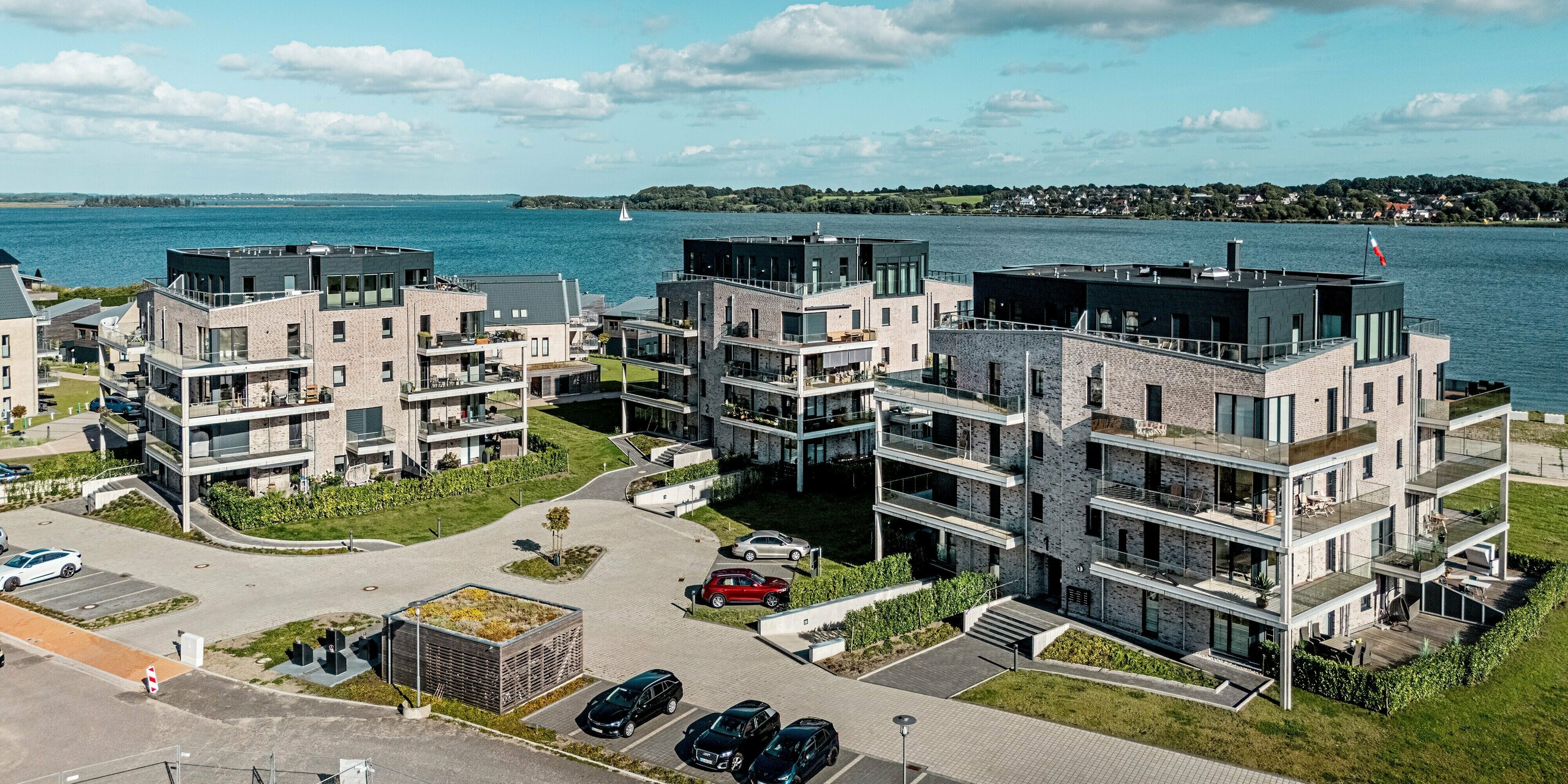 Das Luftbild zeigt die direkt an der Küste gelegene Wohnhausanlage „Hafen zur Freiheit“ in Schleswig, die aus mehreren modernen Gebäuden mit heller Klinkerfassade und markanten Akzenten in PREFA Siding.X in der Farbe P.10 Anthrazit besteht. Die strukturierten Aluminiumpaneele verleihen den oberen Geschossen eine kraftvolle, zeitgemäße Optik und setzen sich klar vom traditionellen Mauerwerk ab. Großzügige Balkone öffnen den Blick auf das Wasser, während die langlebigen und korrosionsbeständigen PREFA-Fassadenelemente in der exponierten Küstenlage optimalen Schutz bieten und die Wertbeständigkeit der Anlage erhöhen.