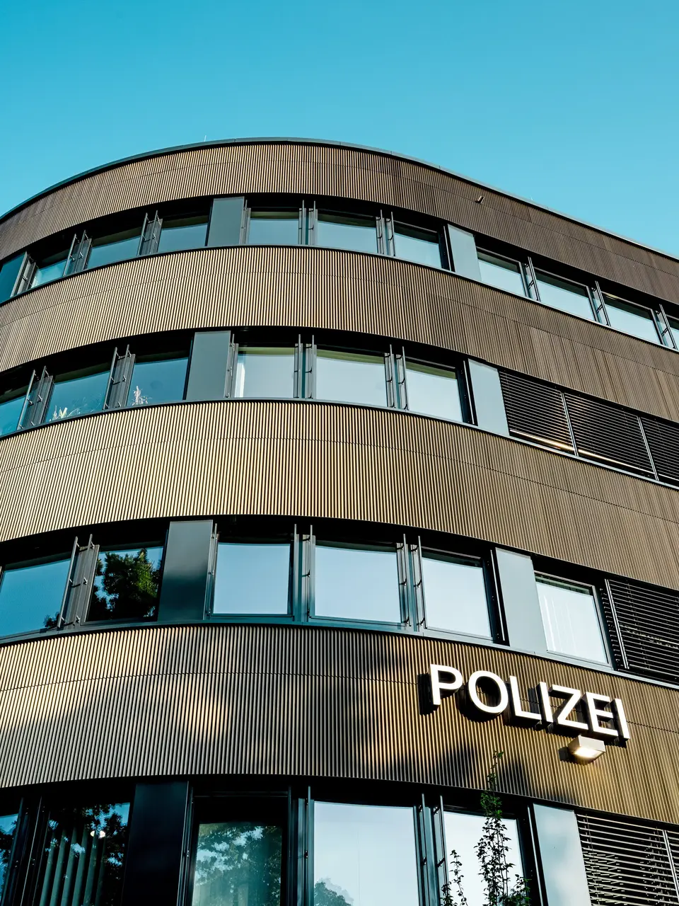 Dramatische Untersicht des geschwungenen Neubaus vom Polizeikommissariat 43 in Hamburg: Die markante PREFA Aluminiumfassade aus eloxierten Strangpressprofilen „Zacke“ in Mittelbronze zieht als architektonischer Eyecatcher alle Blicke auf sich. Die feine, rhythmische Struktur erzeugt dynamische Licht- und Schattenverläufe und betont die fließenden Rundungen des Baukörpers. PREFA Aluminium überzeugt durch hohe Witterungsbeständigkeit, Korrosionsschutz und Langlebigkeit – eine nachhaltige, wartungsarme Lösung für präzise, moderne Fassadenarchitektur.