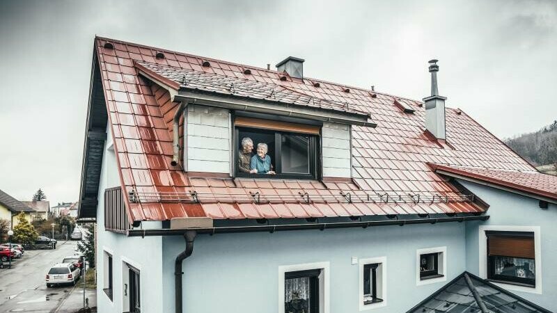 Anna und Heinrich stehen in ihrem Eigenheim - eingedeckt mit der PREFA Dachplatte in der Farbe Ziegelrot - am offenen Fenster und sehen sich glücklich und zufrieden an. 