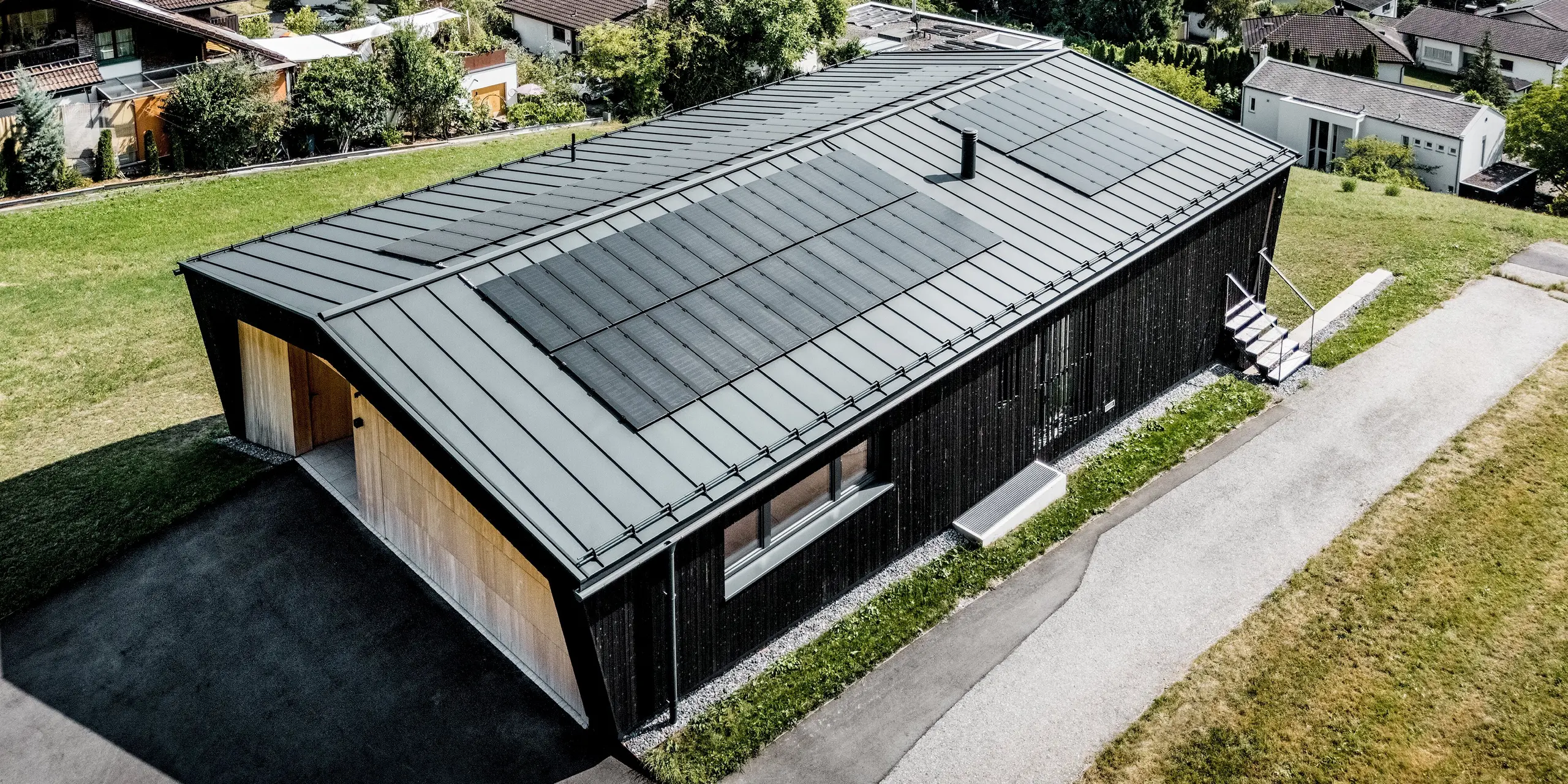 Vogelperspektive auf das Einfamilienhaus in Röthis: Großflächige Solarmodule PREFALZ fügen sich harmonisch in das PREFALZ Satteldach in P.10 Anthrazit ein. Die klaren Stehfalzlinien betonen die moderne Dachgestaltung, während die leichte, langlebige und witterungsbeständige Aluminiumdeckung zuverlässigen Schutz bietet. Die dunkle Holzfassade setzt einen spannenden Kontrast zur hellen Holzverkleidung des Eingangsbereichs.