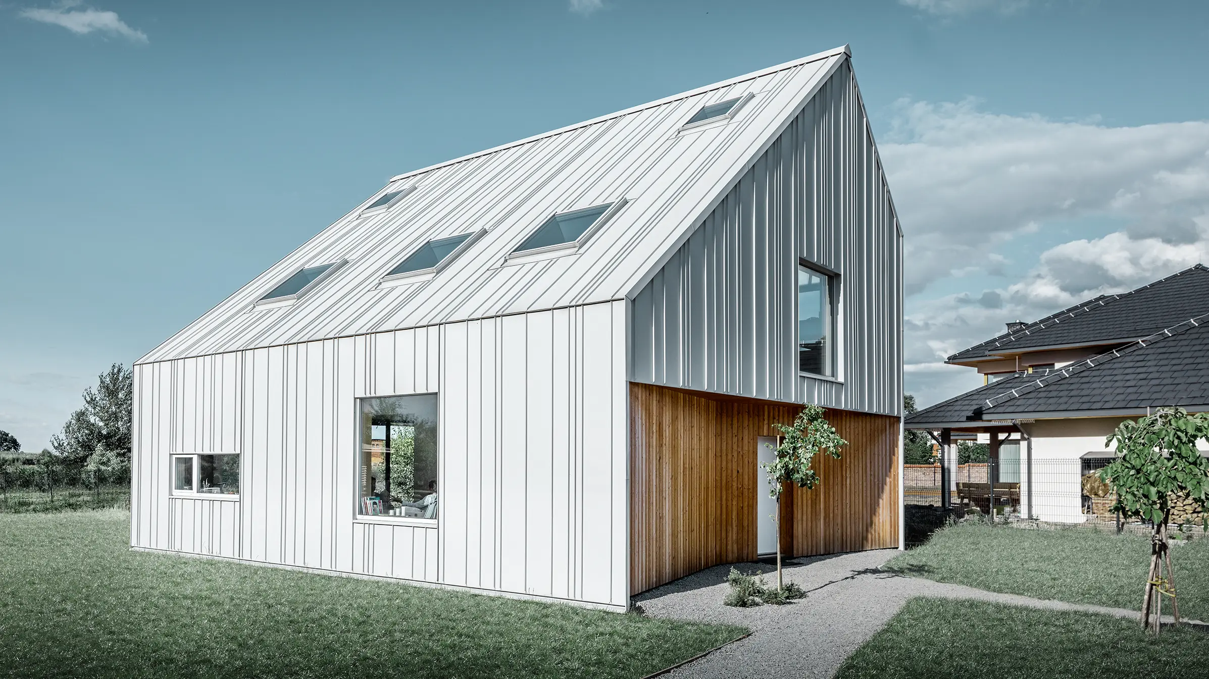 Modern geschnittenes Einfamilienhaus mit Satteldach, die komplette Außenhaut ist mit PREFALZ in Prefaweiß verkleidet. Dabei ziehen sich die unterschiedlich breiten Scharen vom Dach weiter über die Fassade. Im Dach sind mehrere Dachfenster verbaut.