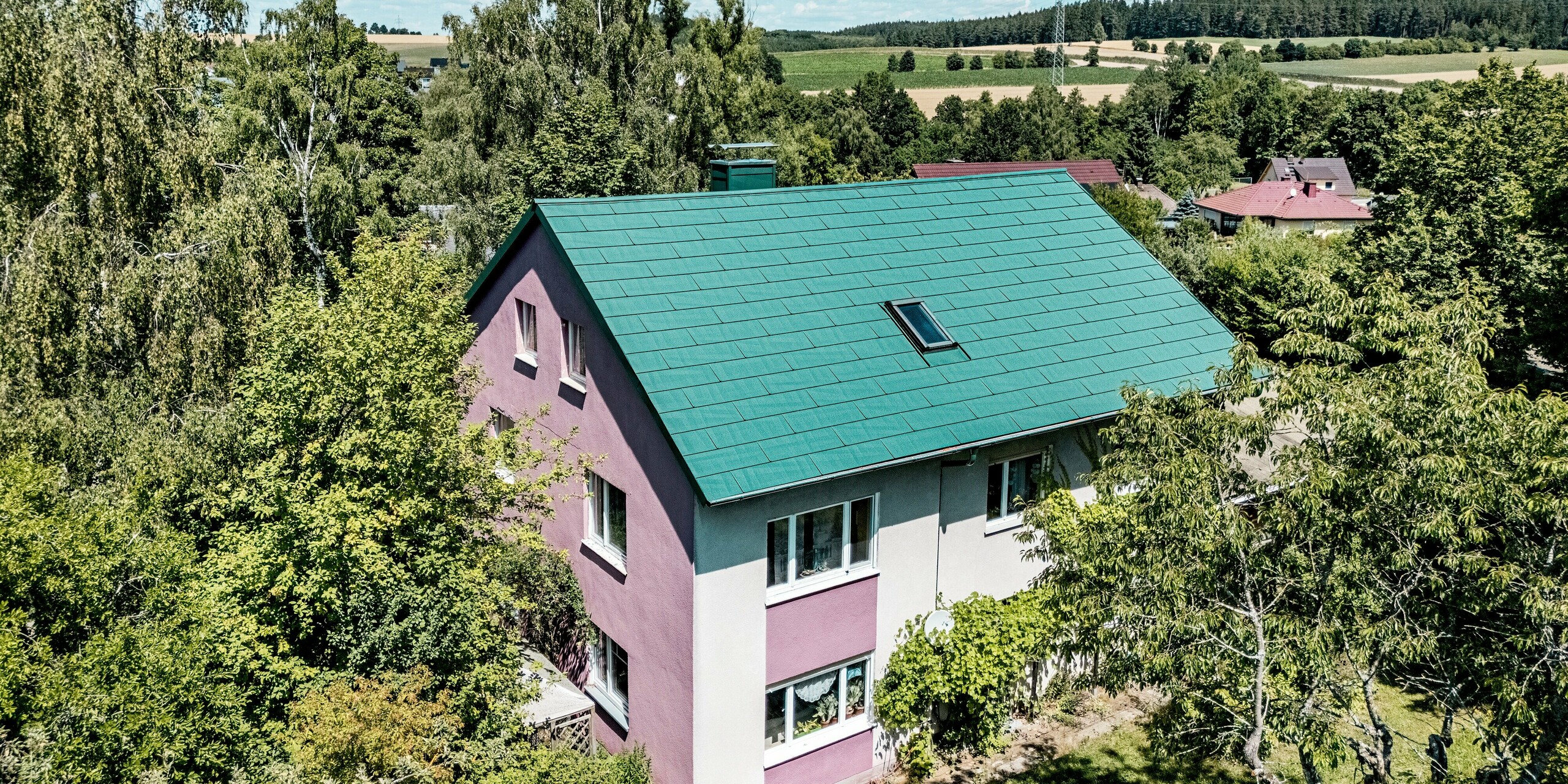 Sanierung eines Einfamilienhauses in Schwarzenbach-Martinlamitz mit PREFA Dachpaneel FX.12 in P.10 Moosgrün. Das Bild zeigt das modernisierte Aluminiumdach in kräftigem Moosgrün, das durch seine klare Linienführung und strukturierte Oberfläche auffällt. Der Kontrast zur violett-weißen Fassade verleiht dem Gebäude einen einzigartigen Charakter. Eingebettet in die grüne Landschaft wirkt das Dach modern und dennoch naturnah. Langlebig, sturmsicher und wartungsfrei – das PREFA Dach vereint Funktionalität mit stilvollem Design.