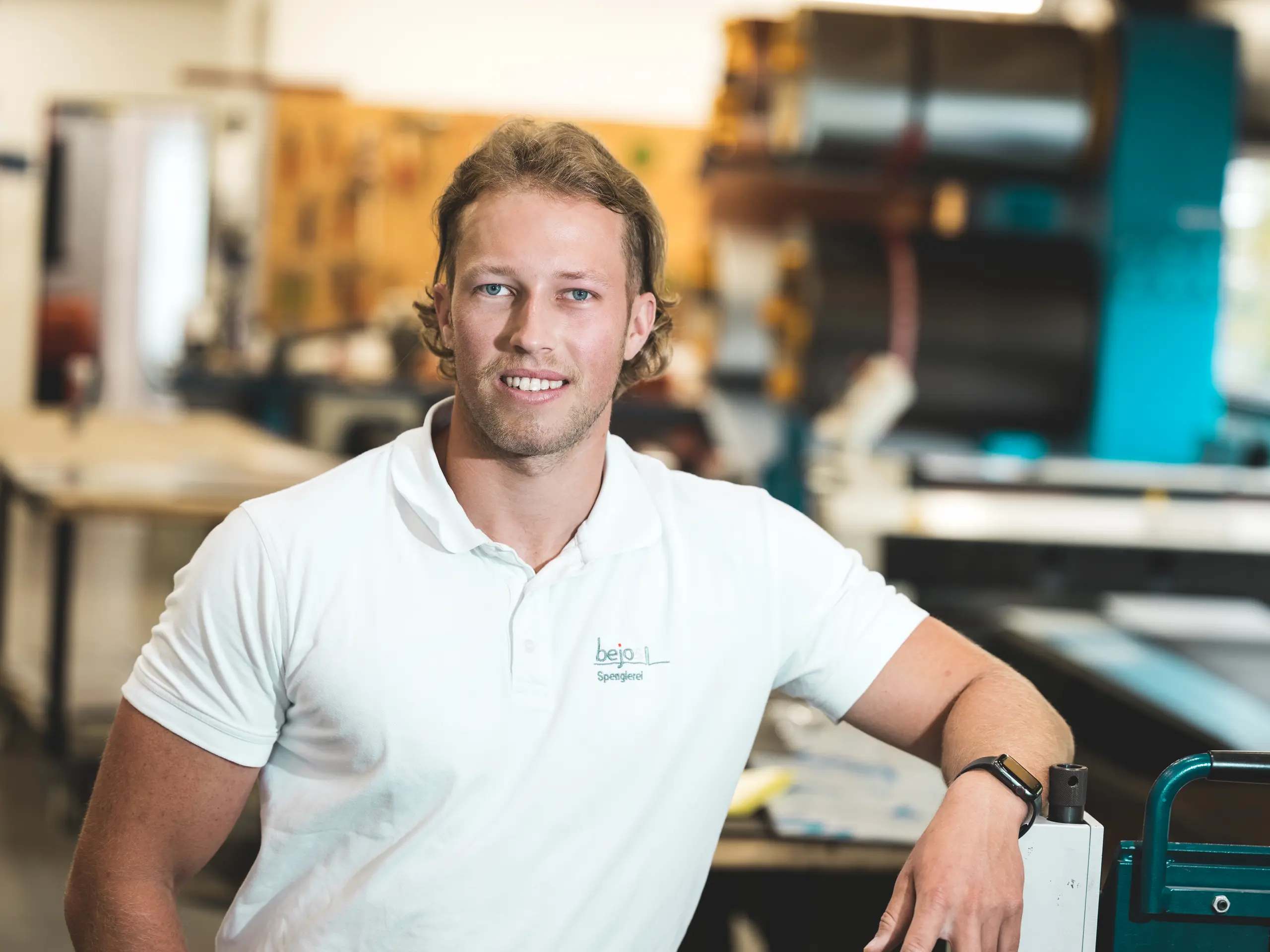 Porträt von Spenglermeister Jakob Berchtold von der Vorarlberger Firma Bejos in seiner Werkstatt. Der Handwerker lehnt entspannt an einer Maschine und blickt lächelnd in die Kamera. Beim Haus Immi realisierte er das integrierte PREFA Solardach aus Solardachplatten und Dachplatten R.16 in P.10 Schwarz – robust, montagefreundlich und optisch harmonisch.