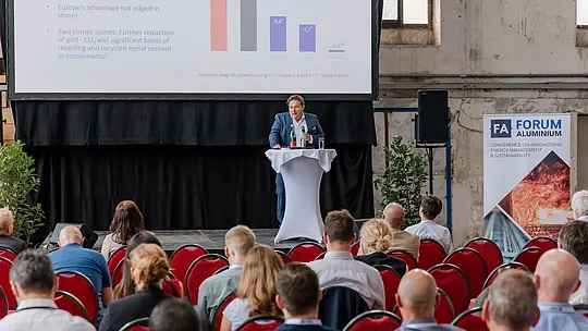 Konferenzszene beim Forum Aluminium im industriellen Ambiente: Im Mittelpunkt steht eine Bühne mit großformatiger Präsentationsleinwand, die Diagramme zu CO₂-Reduktion, Recycling und Markttrends zeigt. Ein Vortragender am Stehtisch erläutert die Inhalte vor einem aufmerksam sitzenden Fachpublikum auf roten Stühlen. Rechts zieht ein „FA Forum Aluminium“-Banner mit Fokus auf Innovation, Energie und Nachhaltigkeit die Blicke auf sich. Die Kombination aus historischer Industriearchitektur und moderner Präsentation unterstreicht die Dynamik und Zukunftsorientierung der Veranstaltung.