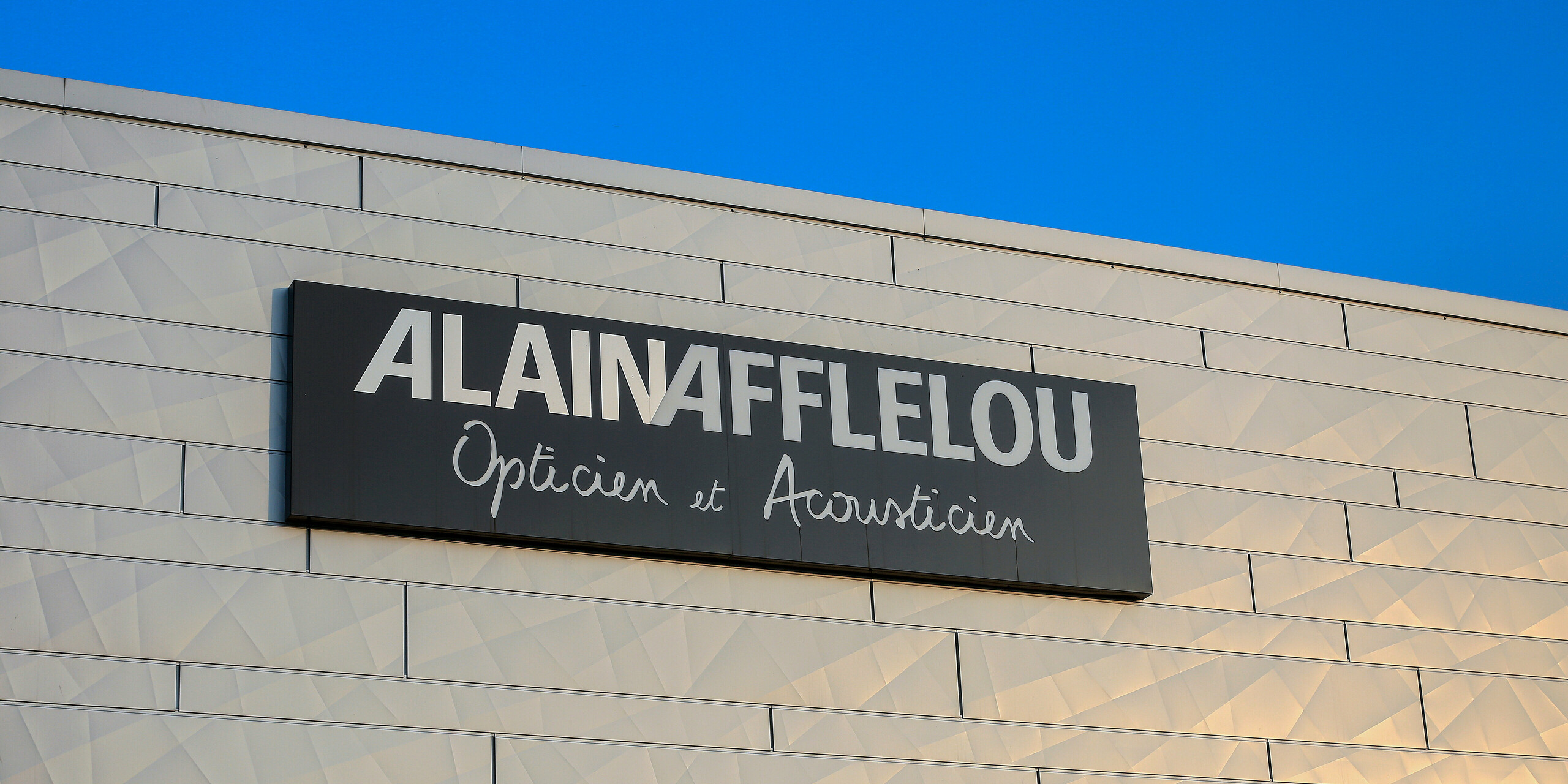 Nahaufnahme des Firmenschilds AFFLELOU am Gebäude des Optik- und Akustik-Shops in Auray - rund um das Logo sind silberschimmernde Fassadenpaneele PREFA Siding.X zu sehen.