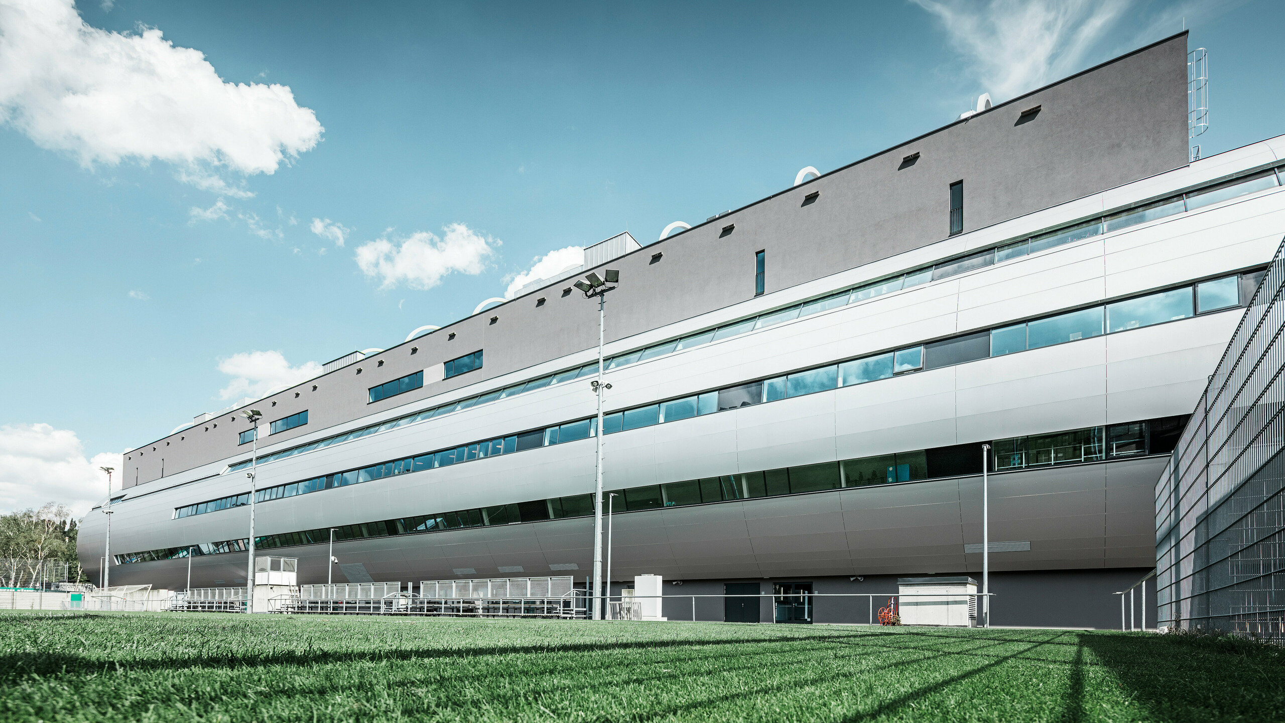 Das Allianz Stadion von SK Rapid Wien, abgebildet vom Trainingsplatz, zeigt die glänzende Fassade aus PREFA Aluminium Verbundplatten in Silbermetallic. Die harmonische Verschmelzung von funktionaler Sportstätte und eleganter Architektur wird durch die Fensterreihen betont. Die grüne Wiese des Trainingsplatzes am unteren Bildrand wird durch die Betreuerbänke und den Zaun abgegrenzt. Am Zaus sind zudem hohe Flutlichtmasten zu erkennen, die eine Ausübung des Trainings zu jeder Tageszeit ermöglichen. Der blaue Himmel komplementiert das professionelle Foto.
