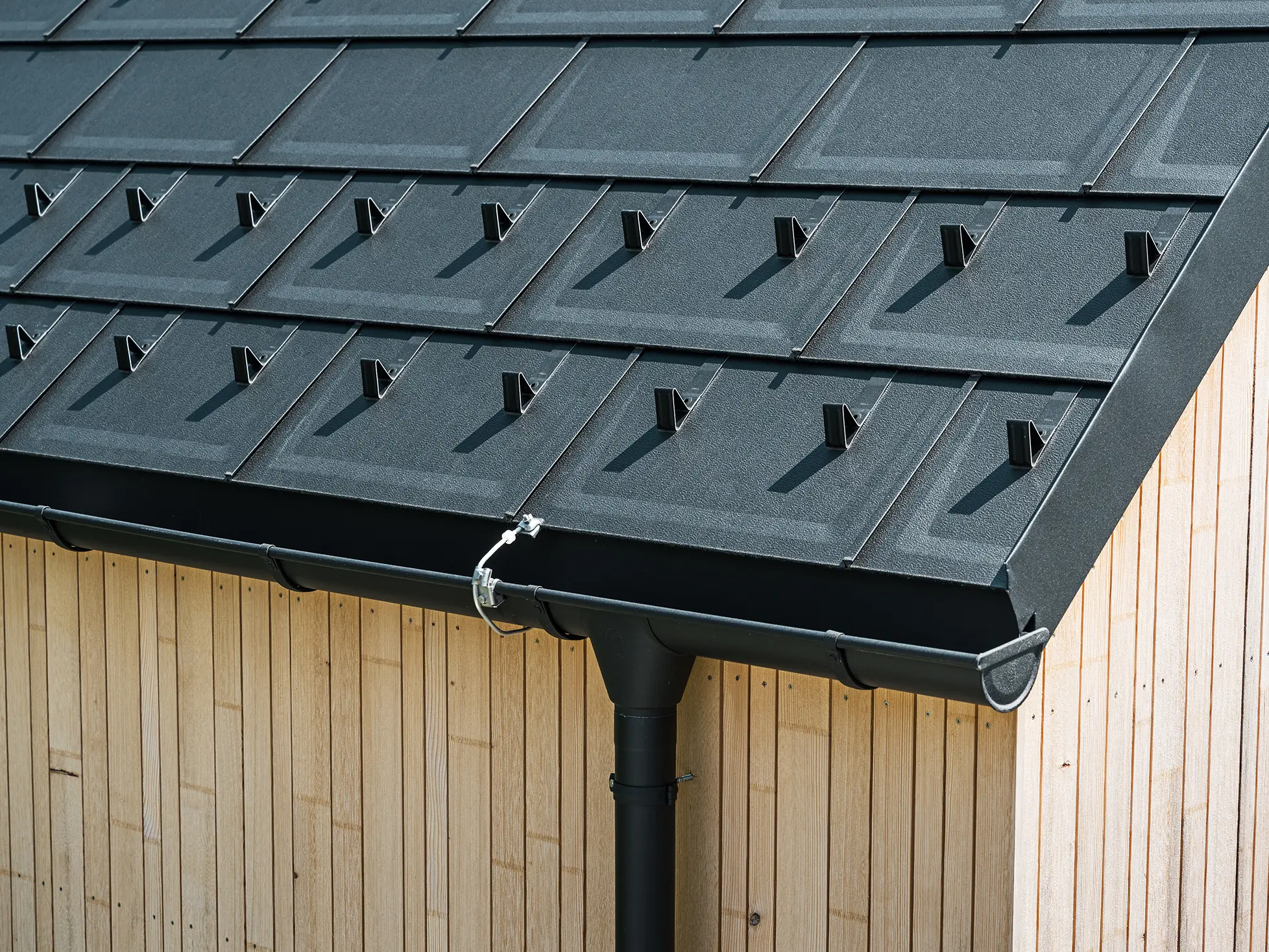Nahaufnahme der PREFA Dachplatte R.16 mit Schneestoppern, Dachrinne und Ablaufrohr in Schwarz P.10 inkl. Blitzschutz und einer senkrechten Holzfassade. Die robusten PREFA Aluminiumprodukte schmücken das Satteldach eines Einfamilienhauses in Lustenau, Vorarlberg.