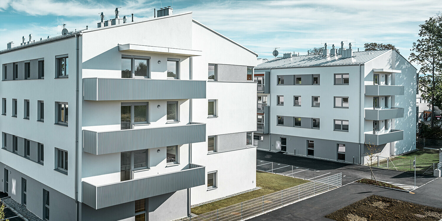 Perspektive aus der Ferne: Die moderne Wohnhausanlage der Glanzstoffsiedlung in St. Pölten präsentiert sich mit einer weißen Fassade und Balkonen, die mit dem PREFA Zackenprofil in Silbermetallic verkleidet sind. Die Aluminiumverkleidung der Balkone bildet einen harmonischen Blickfang in der Umgebung.