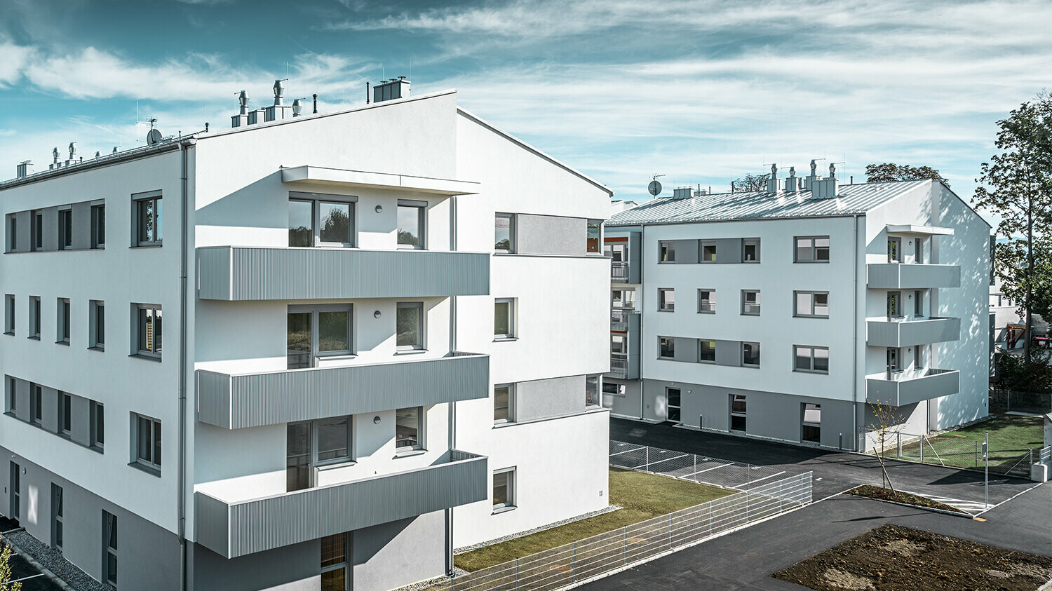 Perspektive aus der Ferne: Die moderne Wohnhausanlage der Glanzstoffsiedlung in St. Pölten präsentiert sich mit einer weißen Fassade und Balkonen, die mit dem PREFA Zackenprofil in Silbermetallic verkleidet sind. Die Aluminiumverkleidung der Balkone bildet einen harmonischen Blickfang in der Umgebung.
