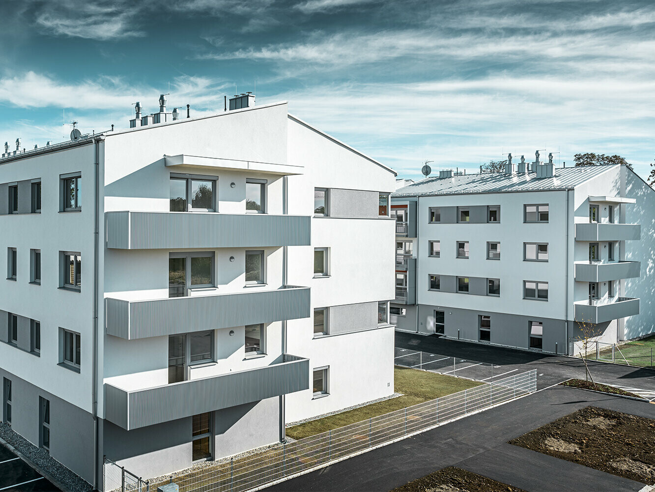 Perspektive aus der Ferne: Die moderne Wohnhausanlage der Glanzstoffsiedlung in St. Pölten präsentiert sich mit einer weißen Fassade und Balkonen, die mit dem PREFA Zackenprofil in Silbermetallic verkleidet sind. Die Aluminiumverkleidung der Balkone bildet einen harmonischen Blickfang in der Umgebung.