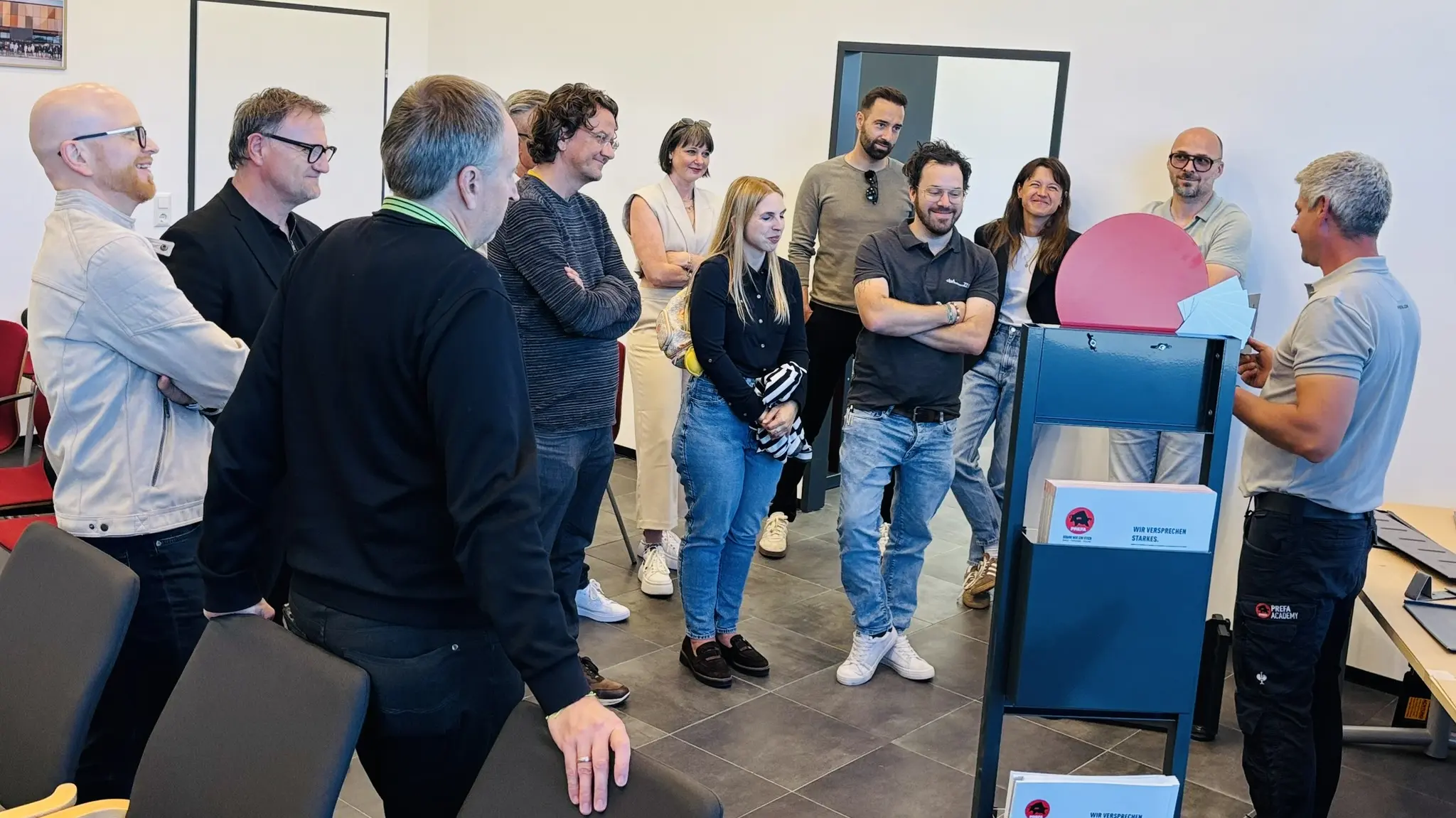 Teilnehmende des PREFA Barcamp-Abends im Showroom St. Veit an der Glan verfolgen aufmerksam eine technische Präsentation zu Aluminium-Dach- und Fassadenlösungen. Eine Person demonstriert ein Werkzeug oder Bauteil, während eine Gruppe von Architektur- und Planungspartner:innen im Halbkreis zuhört. Große Fensterflächen geben den Blick auf außenliegende Solarmodule frei, die den Bezug zu nachhaltigen PREFA Systemlösungen unterstreichen. Die Szene zeigt intensiven fachlichen Austausch, praxisnahe Einblicke und die direkte Vermittlung von Know-how rund um moderne Dach- und Fassadenprojekte.