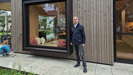 Markus Metzler, PREFA Objektberater, präsentiert während der PREFARENZEN Objekttour in Lustenau die Details eines modernen Wohnhauses mit vertikaler Holzfassade und großzügigem Panoramafenster. Das auskragende, dunkel eingefasste Fensterelement zieht alle Blicke auf sich. Hochwertige PREFA Aluminium-Fensterbänke und Verblechungen setzen präzise Akzente und ergänzen die klare, zeitgemäße Architektur mit langlebiger Funktionalität.