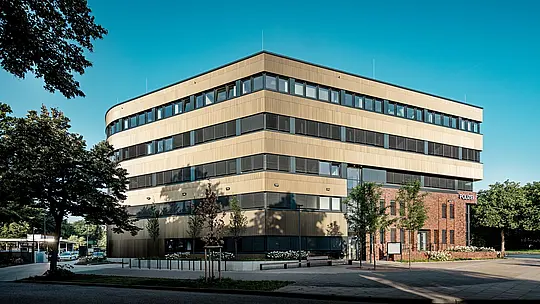 Weitläufige Außenansicht des Polizeikommissariats 43 in Hamburg: Der klar gegliederte Neubau mit geschwungenen Kanten und horizontalen Fensterbändern beeindruckt mit einer rund 1.100 m² großen PREFA Aluminiumfassade aus Strangpressprofilen „Zacke“ in Mittelbronze. Die fein strukturierte Oberfläche wirkt je nach Lichteinfall lebendig und verleiht dem Baukörper Tiefe und Eleganz. Im spannenden Kontrast zum Backsteinbestand entsteht ein markanter Eyecatcher, während PREFA Aluminium durch Langlebigkeit, Korrosionsbeständigkeit und geringen Wartungsaufwand überzeugt.