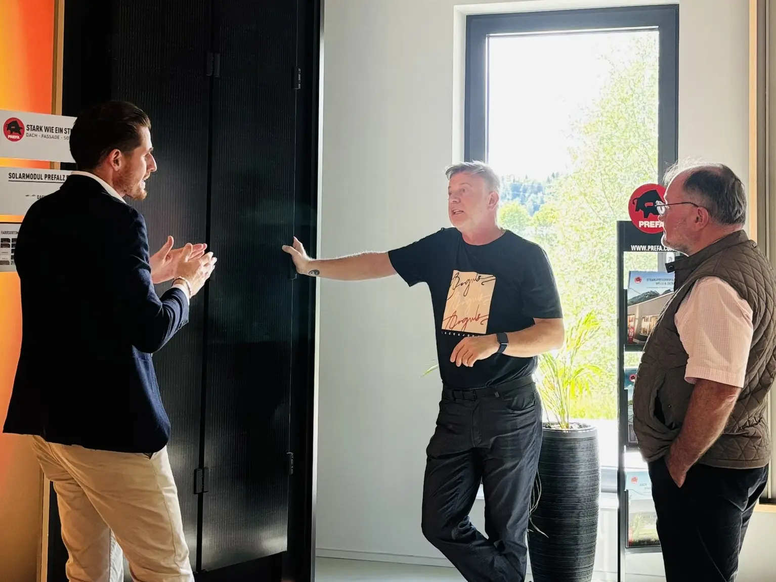 Fachlicher Austausch im PREFA Showroom St. Veit an der Glan: Ralf Stadlober diskutiert mit den Teilnehmenden vor einer großformatigen Musterfläche zu modernen Dach- und Fassadenlösungen mit PREFA Aluminium. Auf der Musterfläche ist das innovative Solarmodul PREFALZ zu sehen, während im Hintergrund Informationen zu Photovoltaik-Lösungen und nachhaltiger Energie sichtbar sind. Die Szene verdeutlicht den praxisnahen Wissenstransfer im Barcamp-Format, bei dem Architektur- und Planungspartner:innen innovative PREFA-Systeme direkt erleben und deren Einsatzmöglichkeiten für Dach und Fassade kennenlernen.