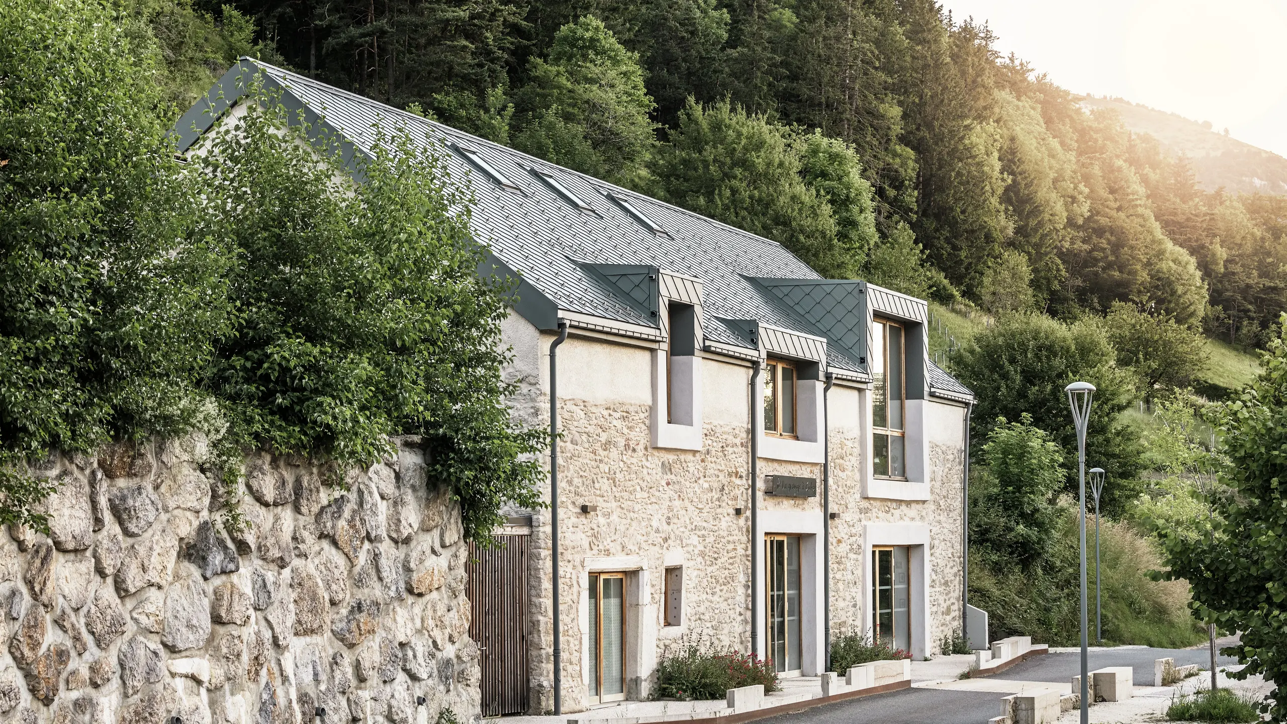 Seitenansicht der restaurierten Scheune „La Grange à Gaby“ in Engins, Frankreich. Das Gebäude vereint traditionelles Steinmauerwerk mit einem modernen PREFA-Aluminiumdach aus 29 × 29 cm großen Rauten. Die sorgfältige Sanierung verbindet historische Bauweisen mit langlebigen Materialien.